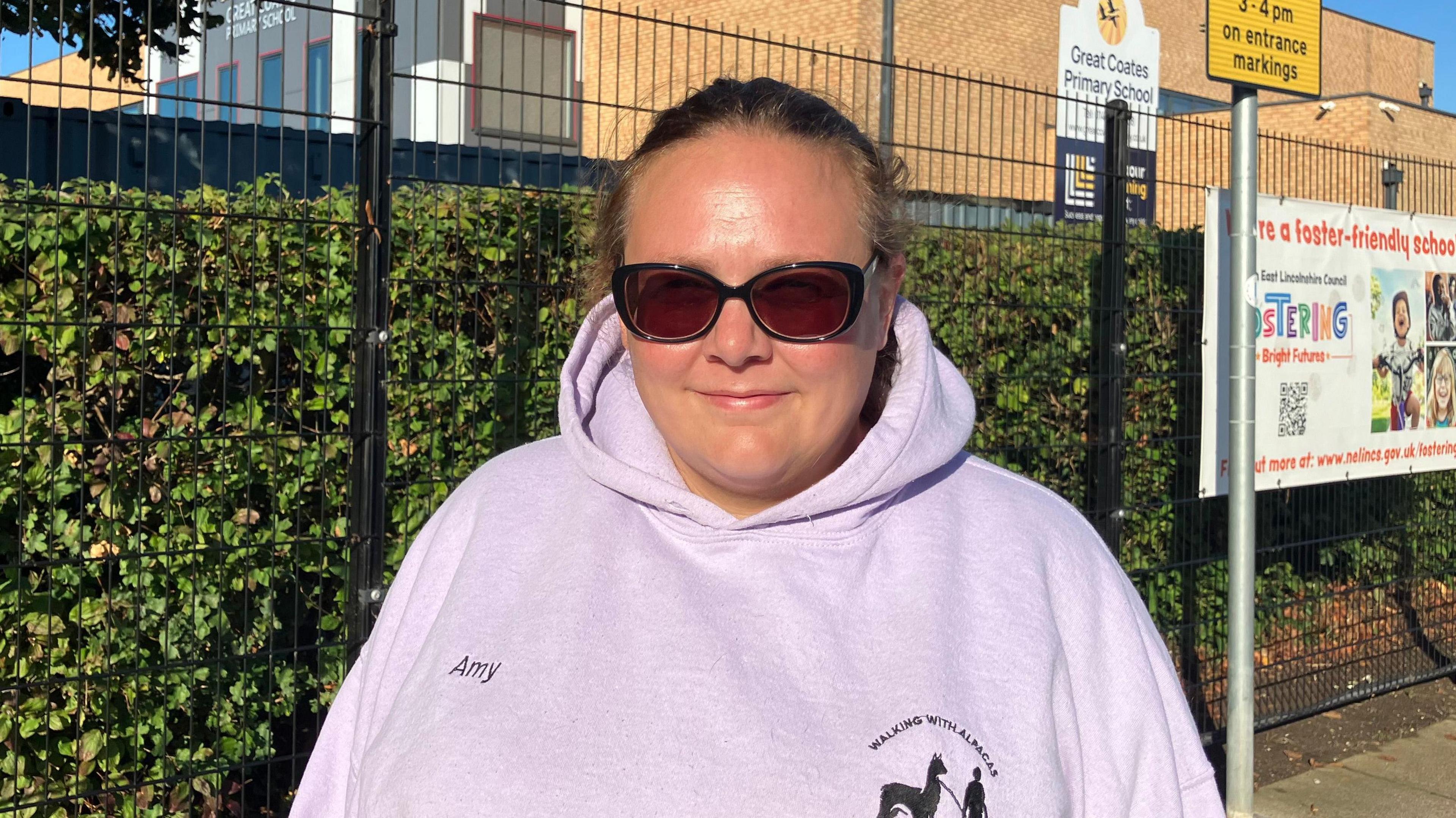 A woman with brown hair, tied back, wearing sunglasses and a light-pink hoodie is standing in front of a modern brick school building, which is surrounded by a black wire fence and green hedge. A yellow parking sign reads: "No stopping Mon - Fri, 8-9am, 3-4 pm on entrance markings". Another sign advertises fostering services.