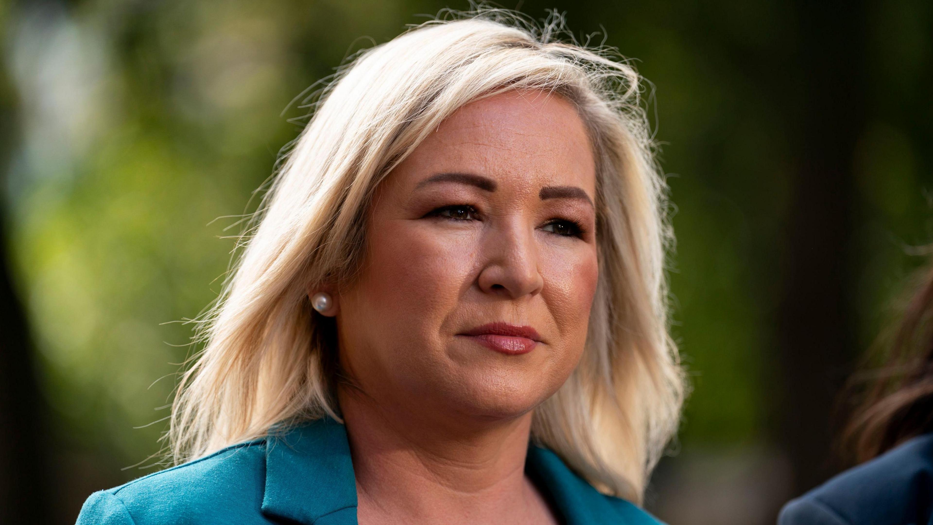 Michelle O'Neill - a woman with blonde, shoulder-length hair looks beyond the camera. She is wearing a green suit jacket. The background is blurred.