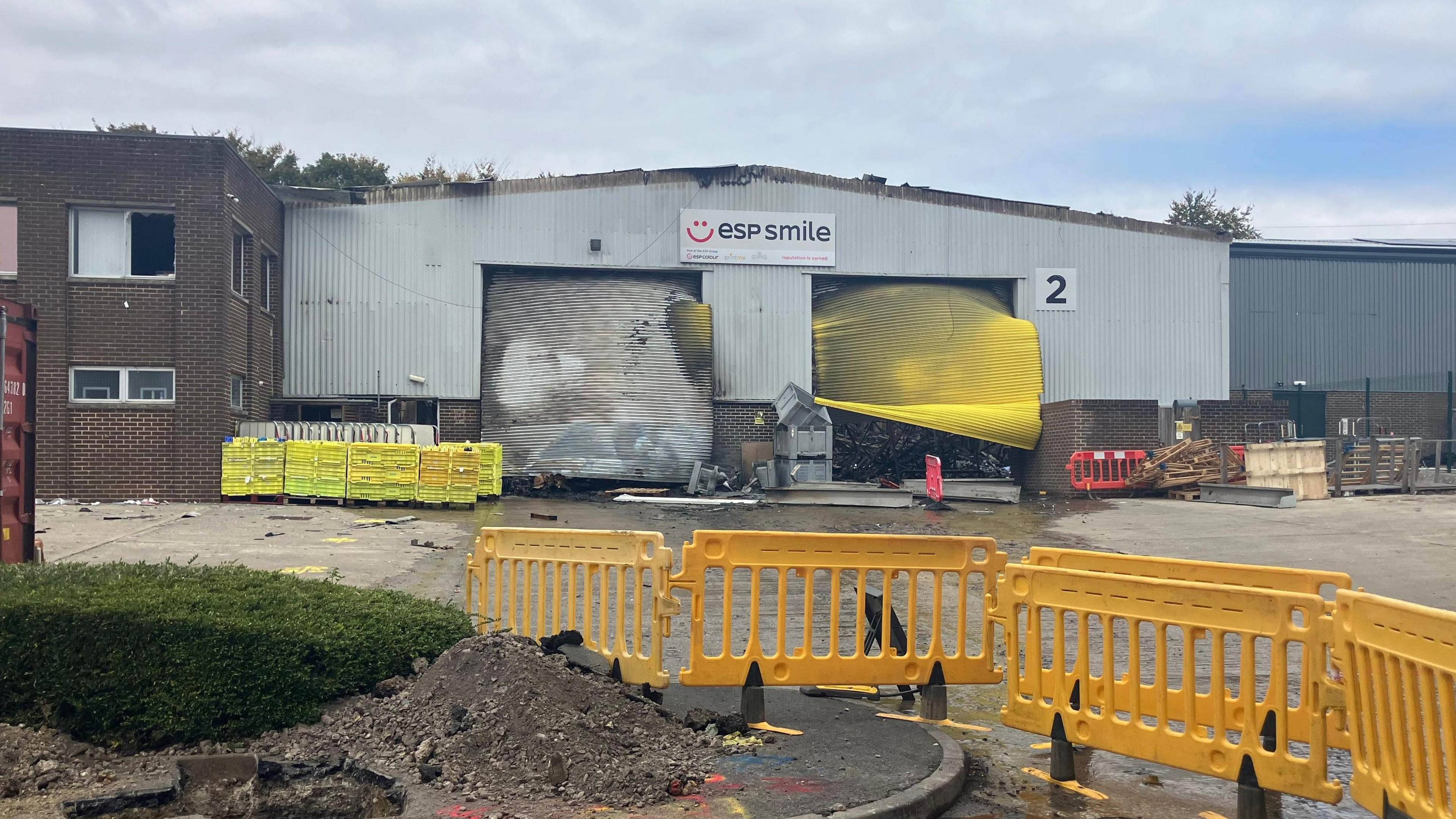 The damage at the scene of an explosion at a warehouse. The grey building has two shutters at the front - one is white and the other is yellow. Both of them are buckled and covered in grey ash. There is a yellow fence cordoning off access to the building. The building has a white sign with a logo for ESP Smile, the business based there.