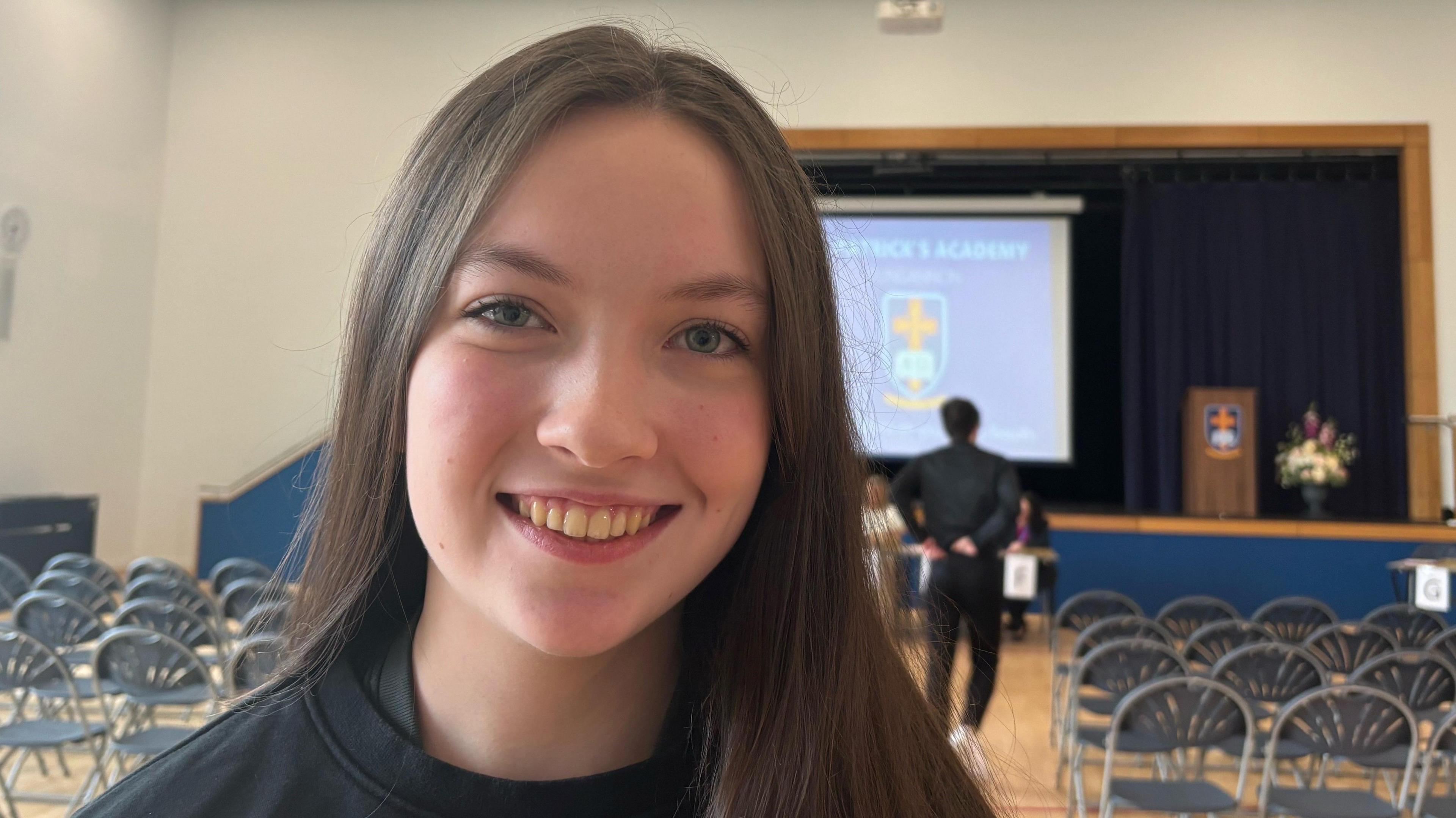 Wide shot of Calla smiling in front of the camera. She has long brown hair and blue eyes. There are school chairs behind her in an assembly hall with a screen with the school logo which is a yellow cross in a white armour. 