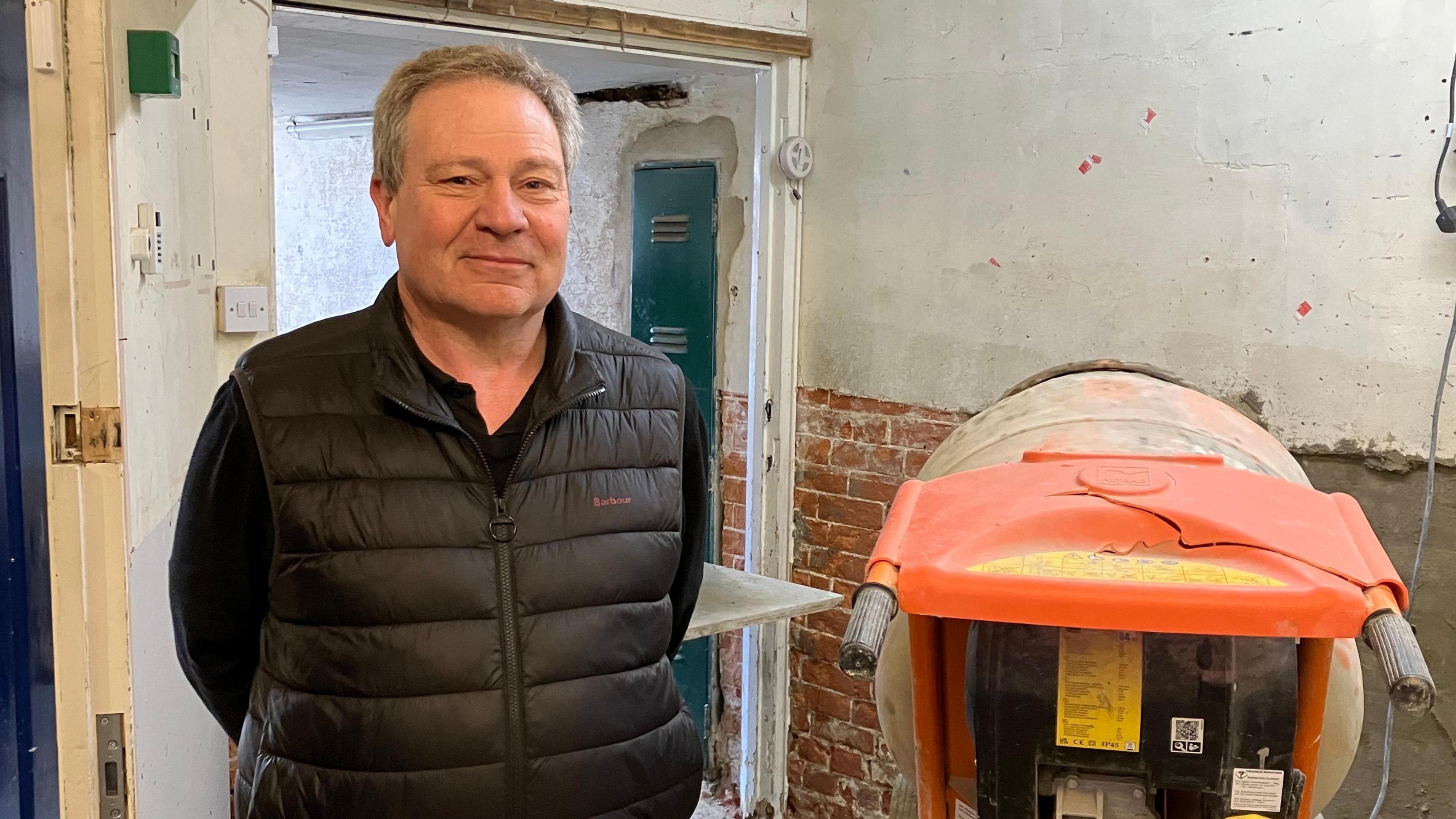 Postmaster wearing black sleeveless puffer jacket standing beside cement mixer 