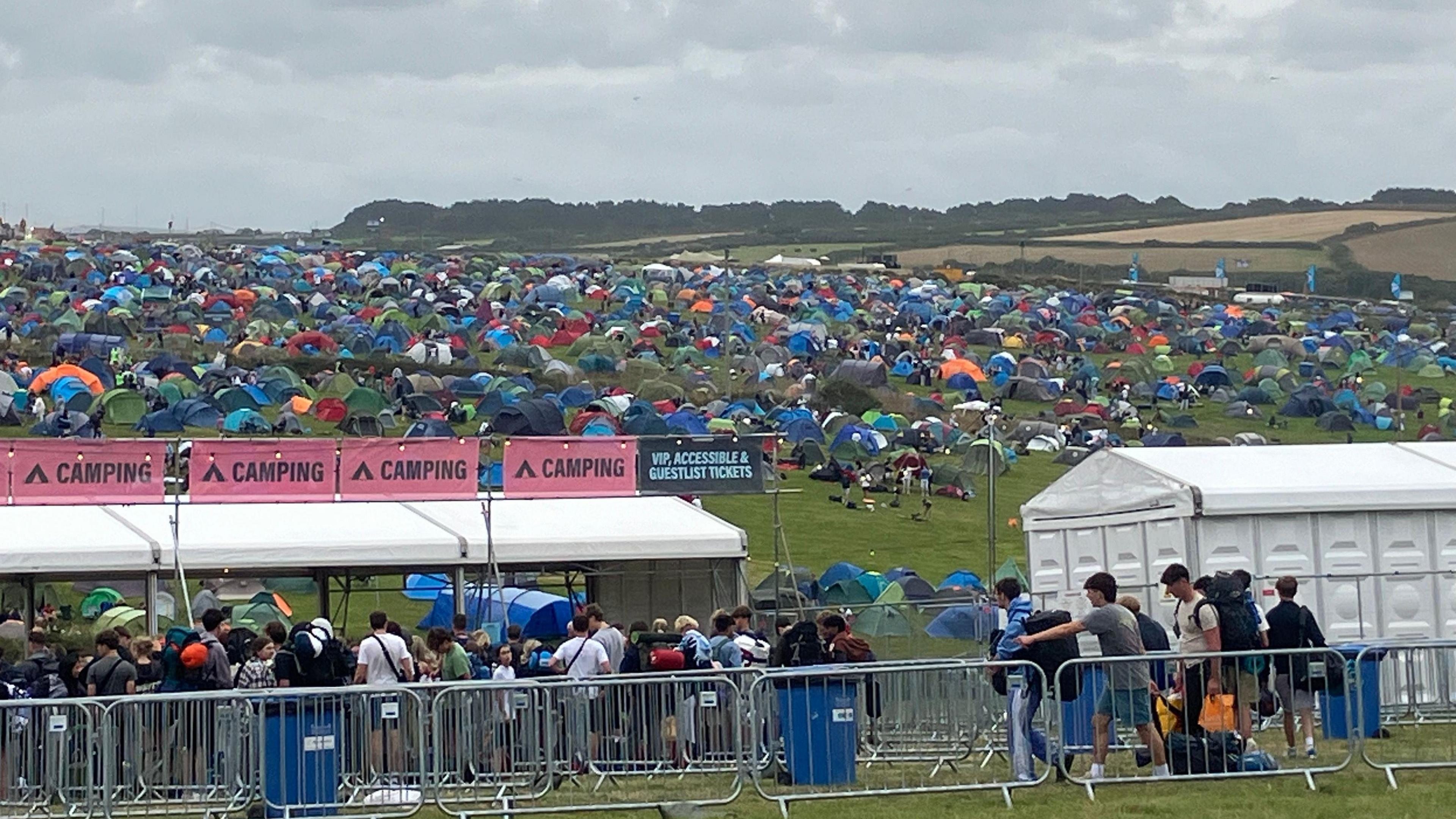 View of Boardmasters Festival campsite. Hundreds of tents are pitched on a sloped green field. In the foreground people are queuing to get access to the campsite at a check-in tent with a white roof