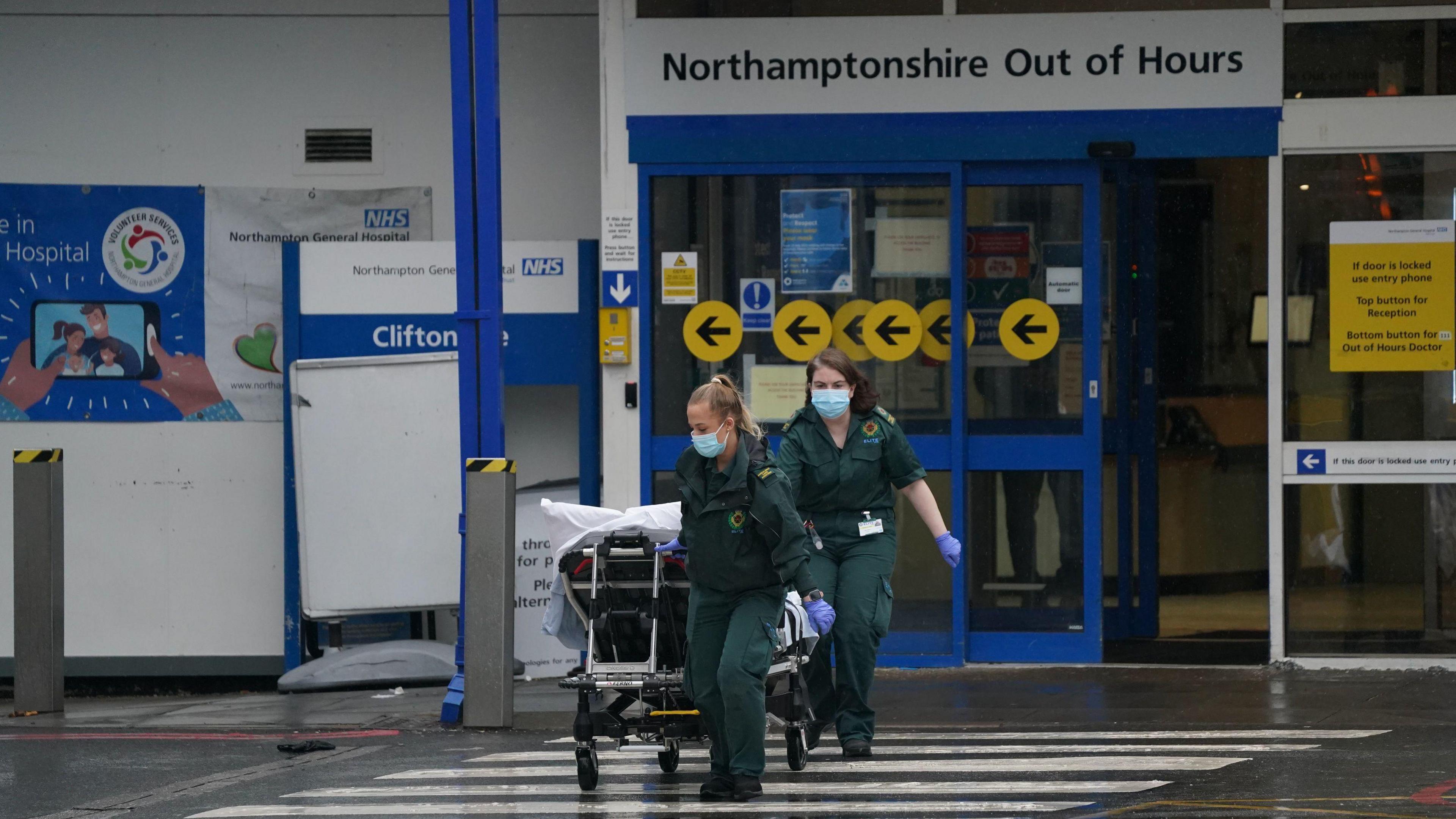 Two ambulance operatives dressed in dark green uniforms with face masks on, wheel a trolley across a zebra crossing out of a hospital reception area. 