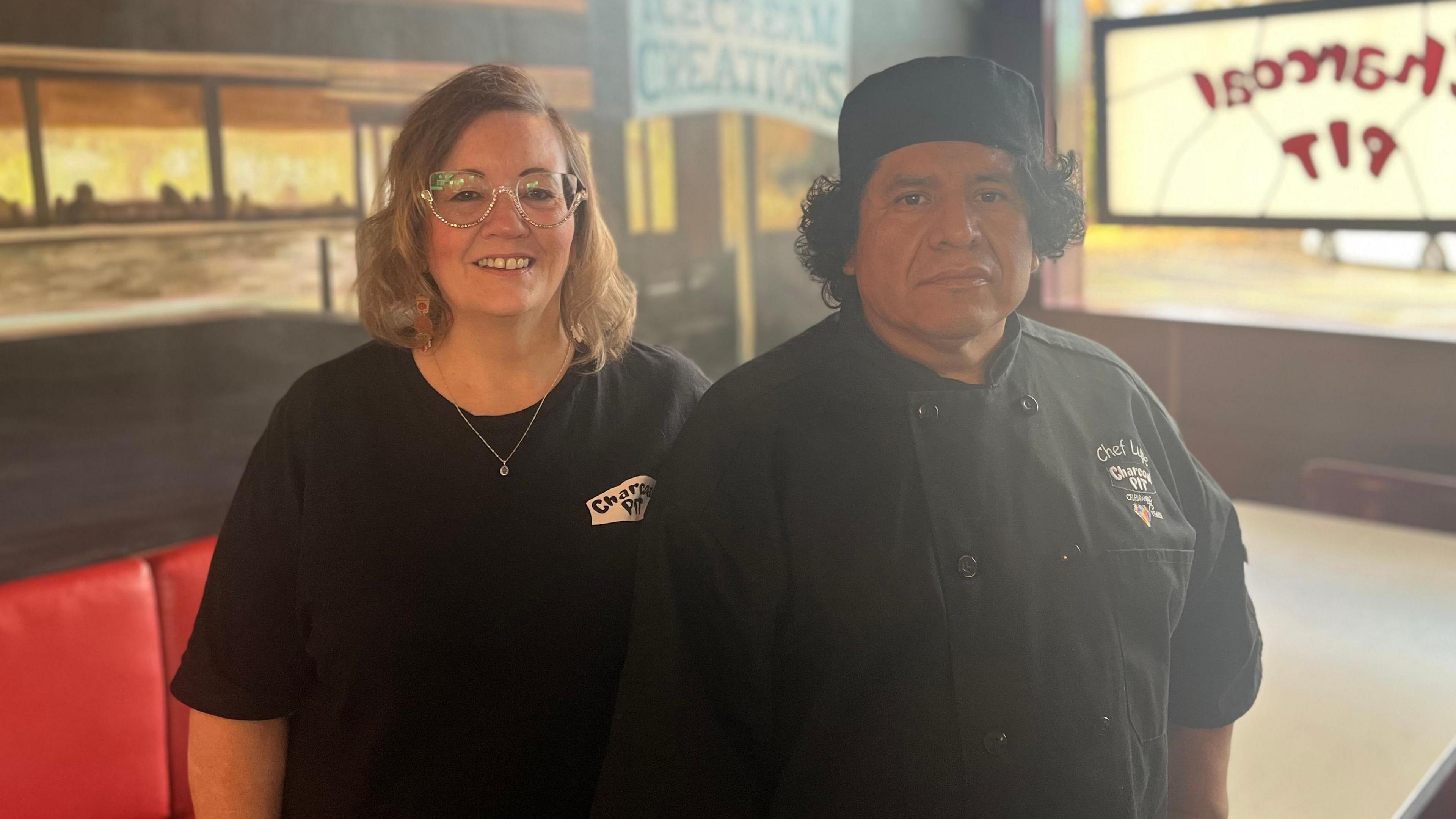 Lupe Avilez and his wife Mary Redden Avilez pictured. He has black long hair and  is wearing a black chef shirt with the company logo and black chef hat. She has shoulder length light coloured hair and is wearing glasses and black t-shirt with the company logo on it.