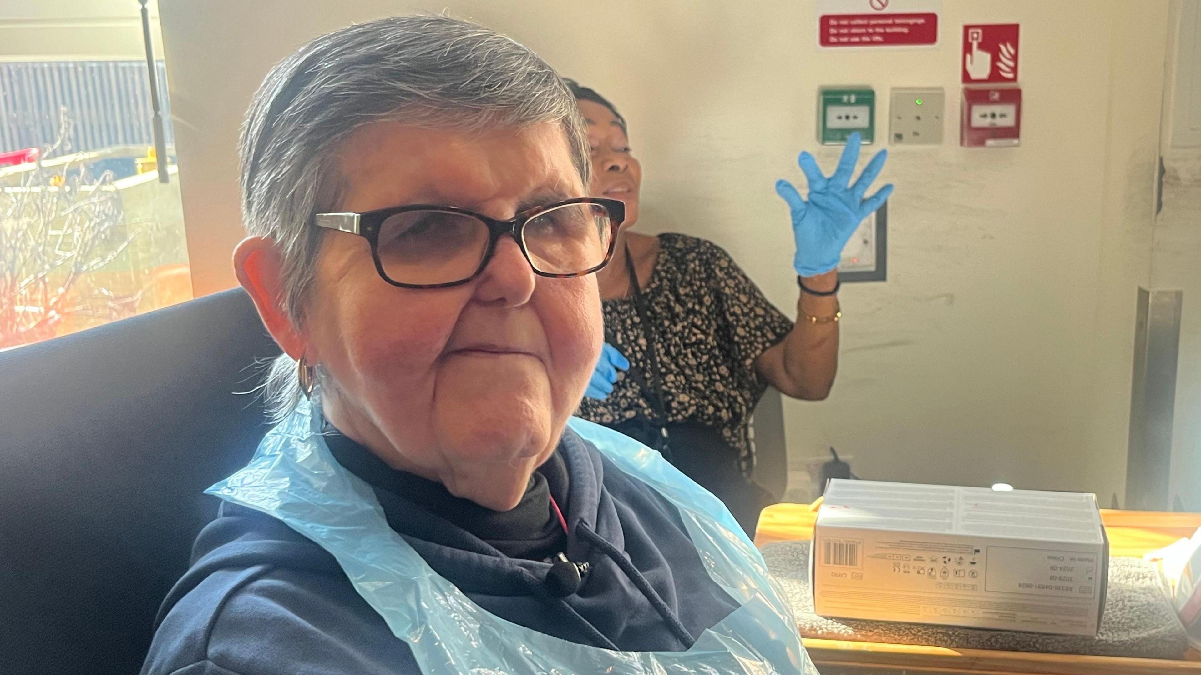 Sharon Elliott smiles at the camera as behind her a member of staff in blue gloves raises her hand