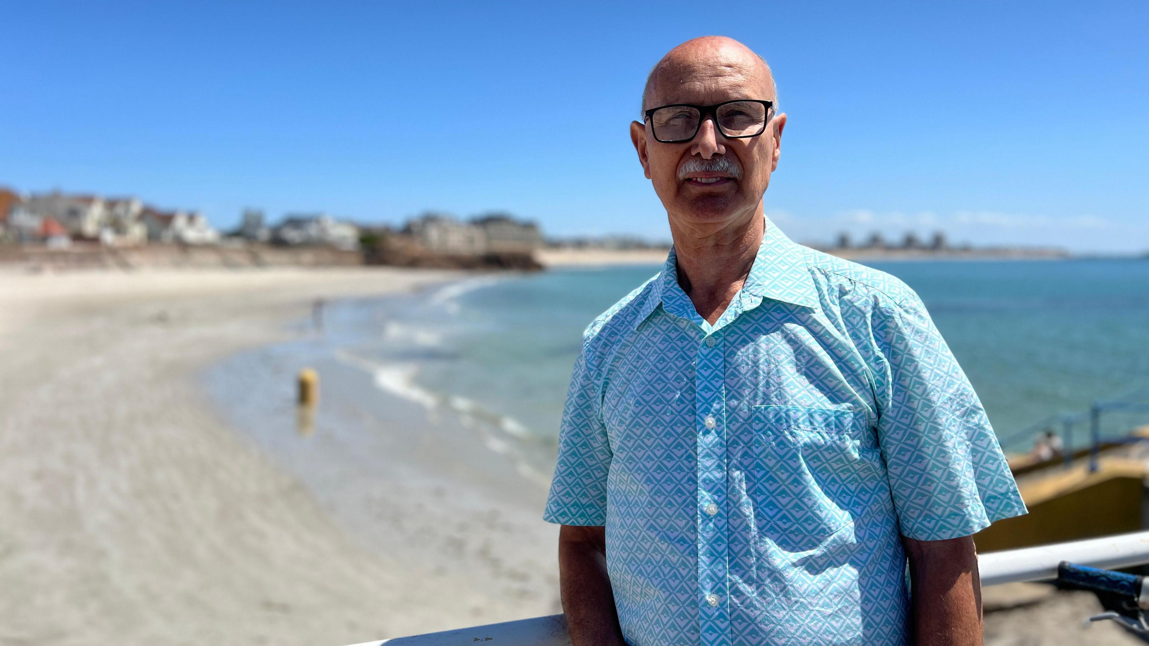 Joao stands on a beachside boardwalk, wearing a light blue patterned short-sleeve shirt. Behind them are a sandy beach, calm ocean waves, and houses along the coastline under a clear blue sky.