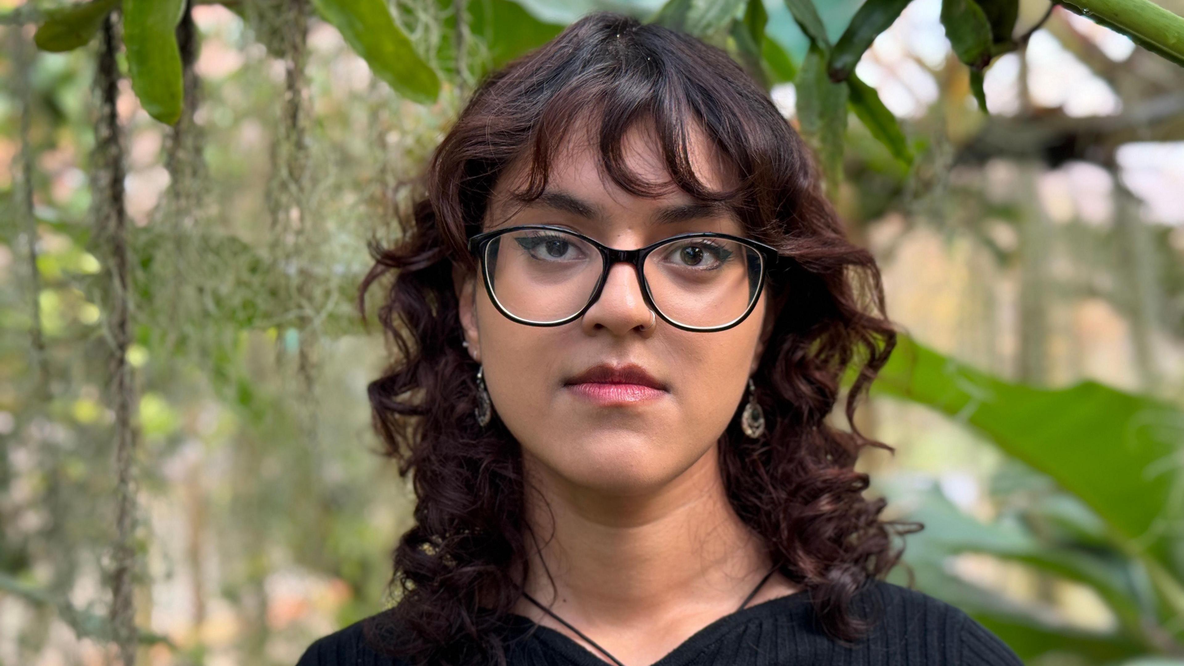 Benika has curly red-brown shoulder-length hair and black glasses. She is wearing a black jumper, and there are green leaves behind her.
