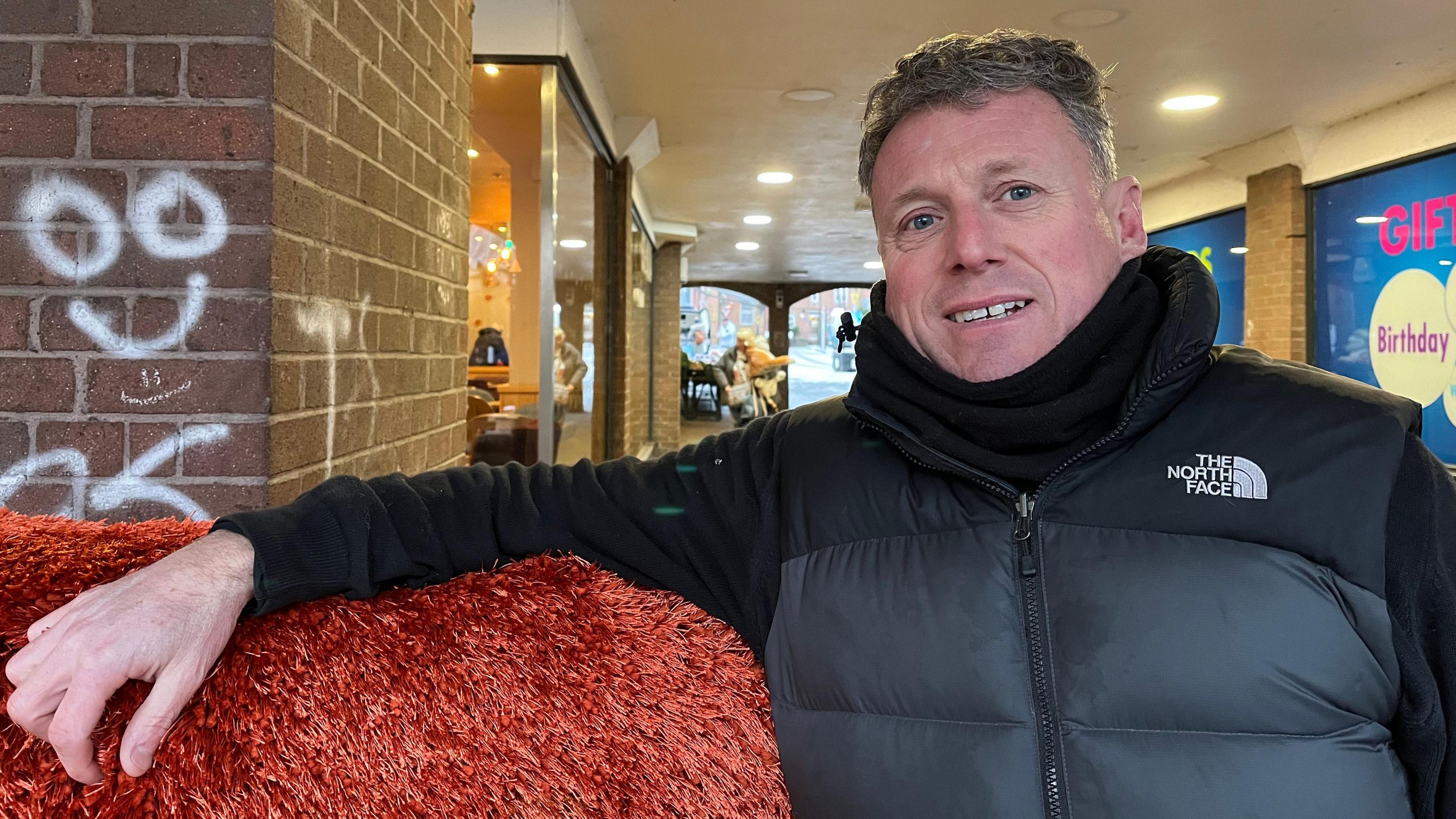 Paul photographed leaning on one of the rugs he sells - it's a bright orange fluffy one. He's wearing a black winter coat, and behind him you can see shops and cafes. 