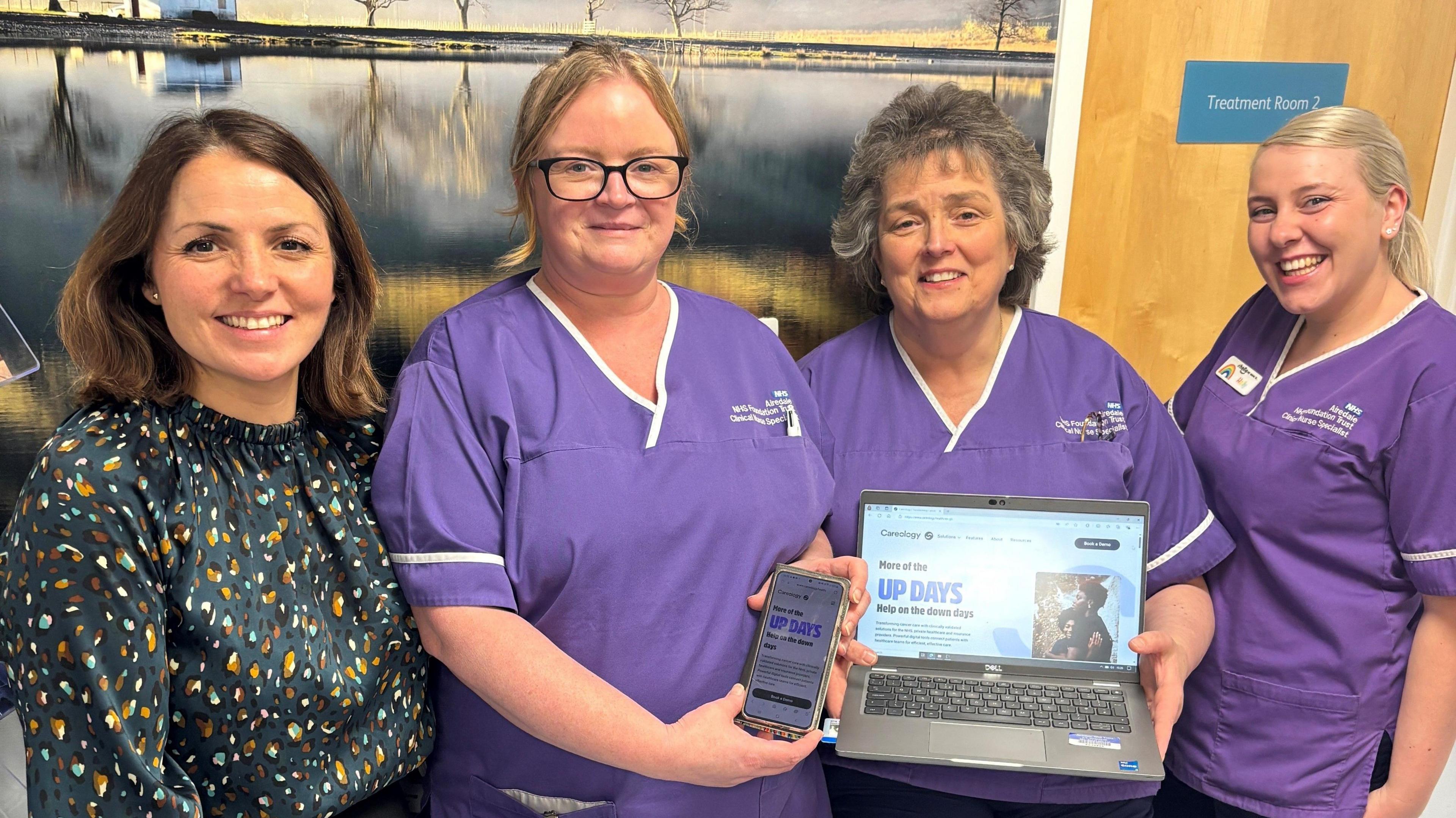 Four women, three in purple nurses' uniforms, holding a mobile phone and laptop displaying the new app.