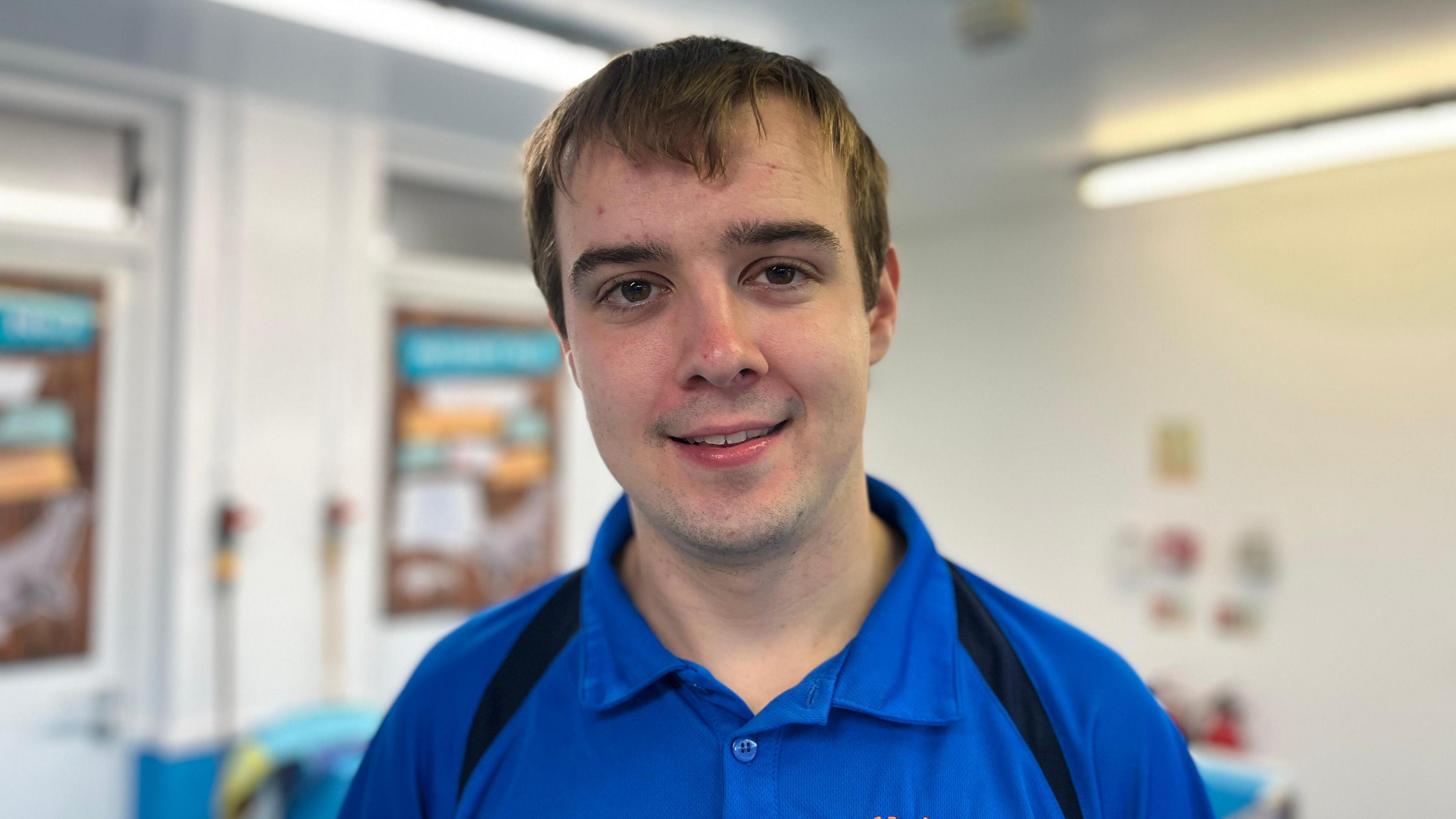 Euan Morrison has brown hair, brown eyes and is smiling at the camera. He is wearing a blue polo short with black stripes on shoulders.