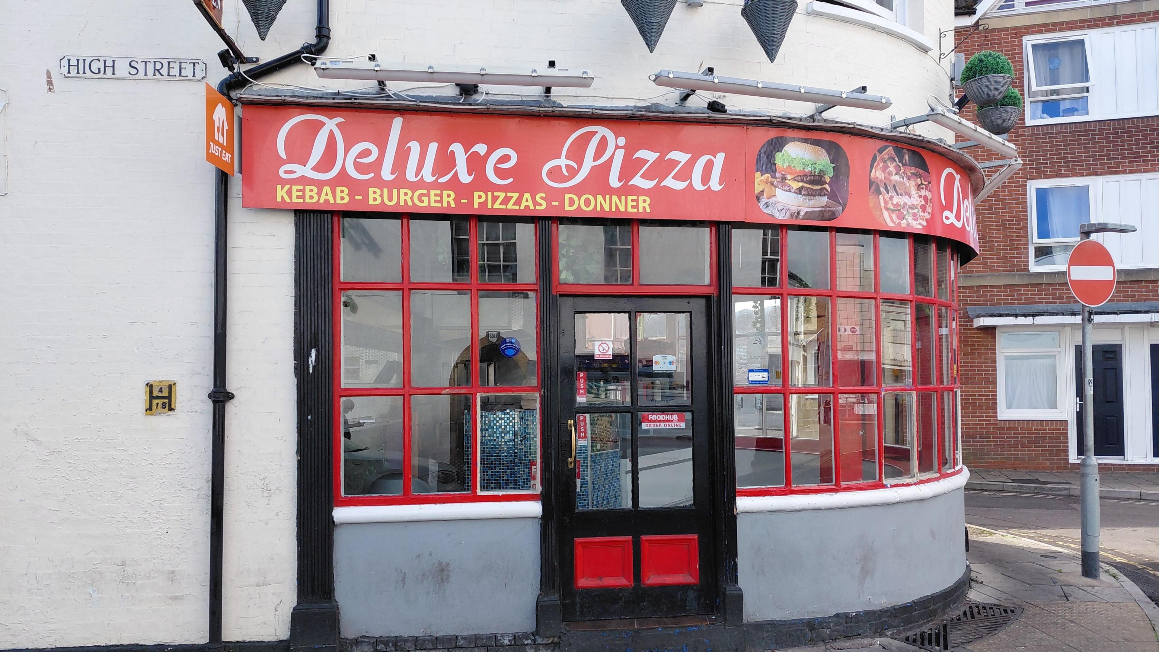 Exterior of Deluxe Pizza, a fast-food restaurant on High Street, featuring red-framed windows and signage advertising kebabs, burgers, pizzas, and doner. The corner building includes food imagery and a prominent red and white sign above the entrance.