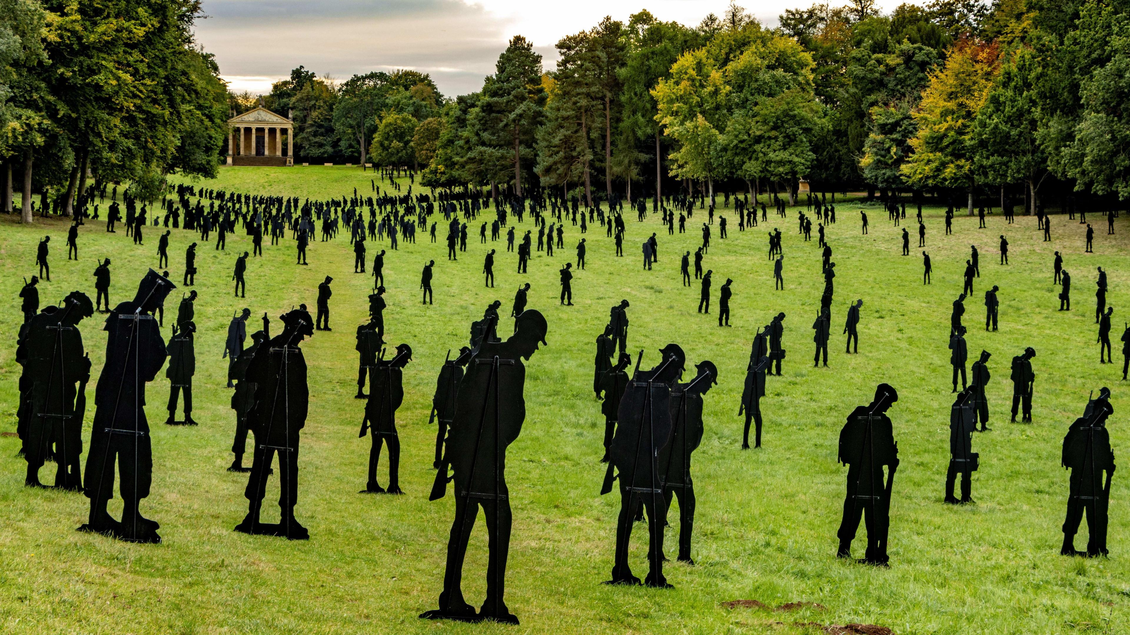 A large number of black silhouettes of servicemen in a garden, showing a Grecian style structure in the distance.  The black silhouettes are placed on green grass and there are lots of trees in the background. 