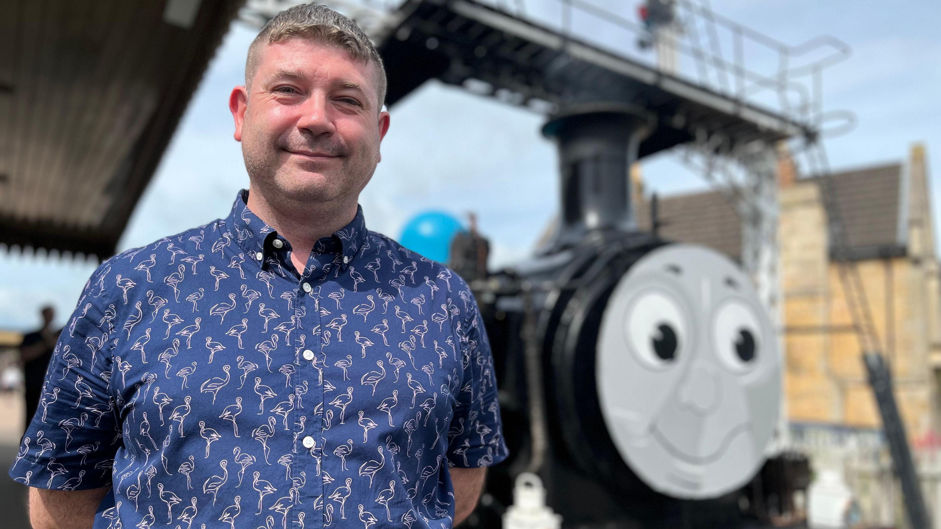 Chris wearing a blue button-down shirt with white flamingos - standing in front of a Thomas train.
