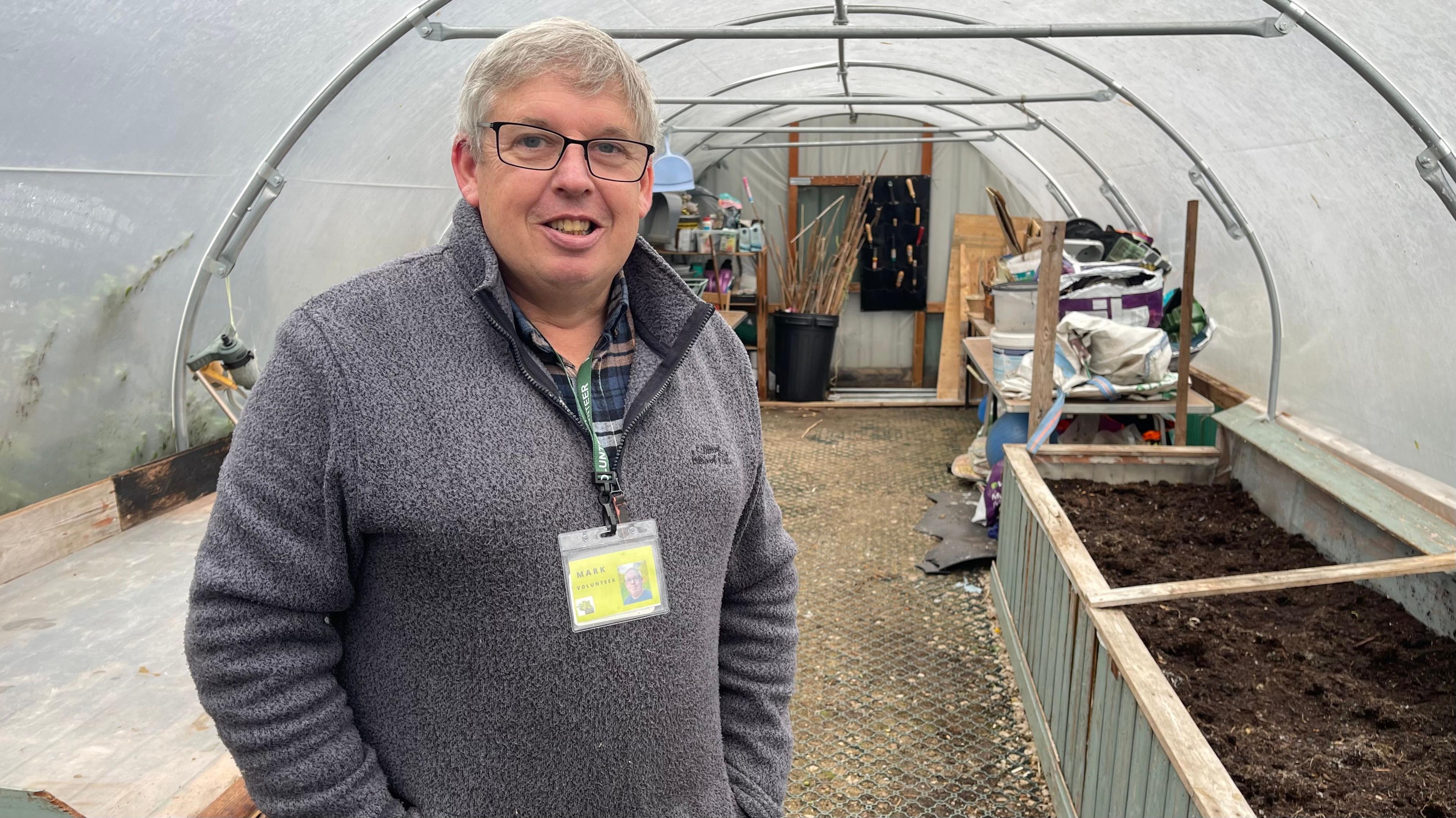 A man stands in a grey 1/4 zipped fleece stands in a garden poly tunnel. Planters are to the right filled with soil, there is a workbench to the left and tools and gardening equipment stands at the end of the tunnel. The man is smiling, with short great hair, glasses and a green lanyard with a yellow ID badge.