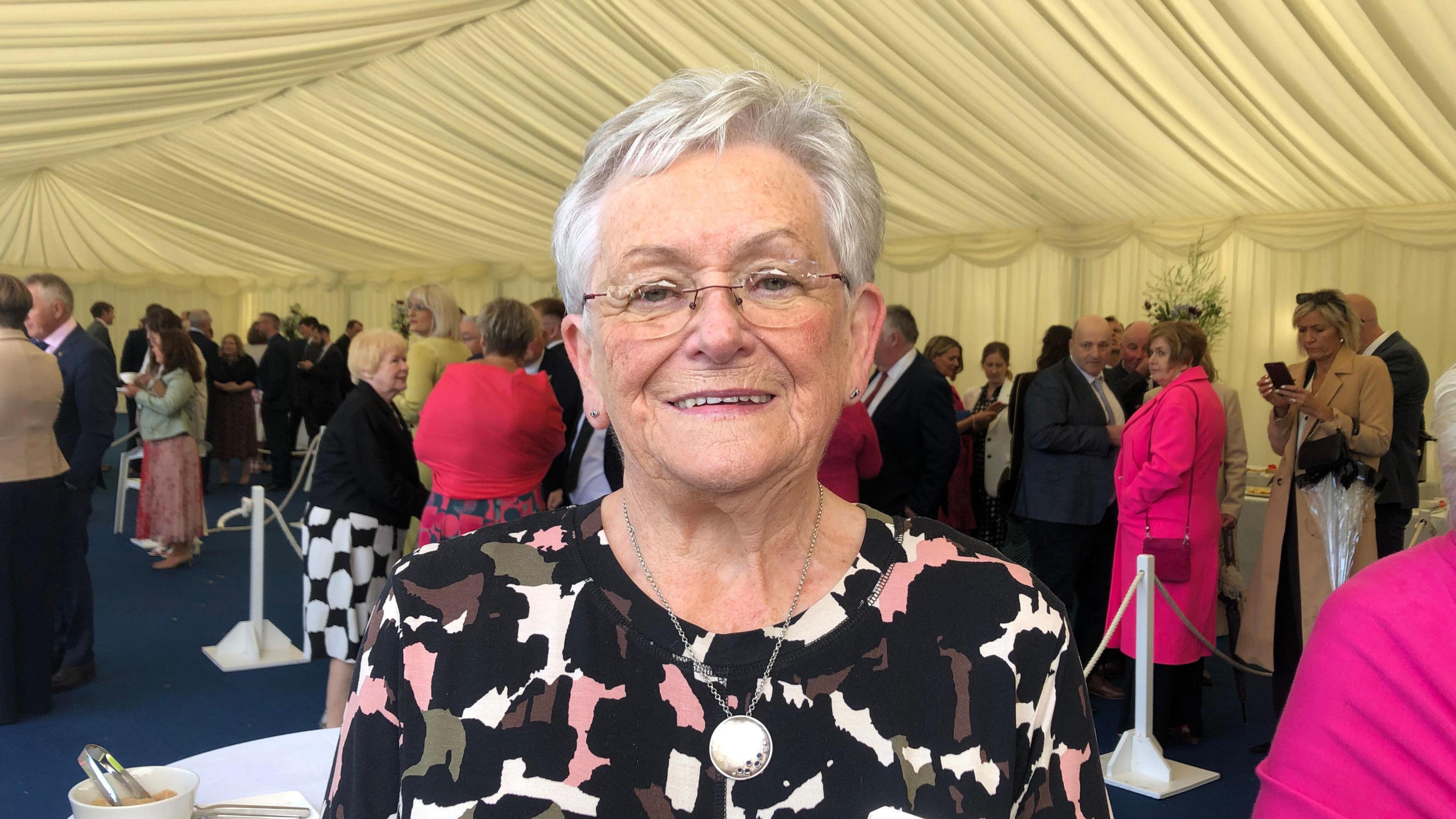 Women with grey hair standing under party tent. She is wearing a pink and green top with a large necklace. 