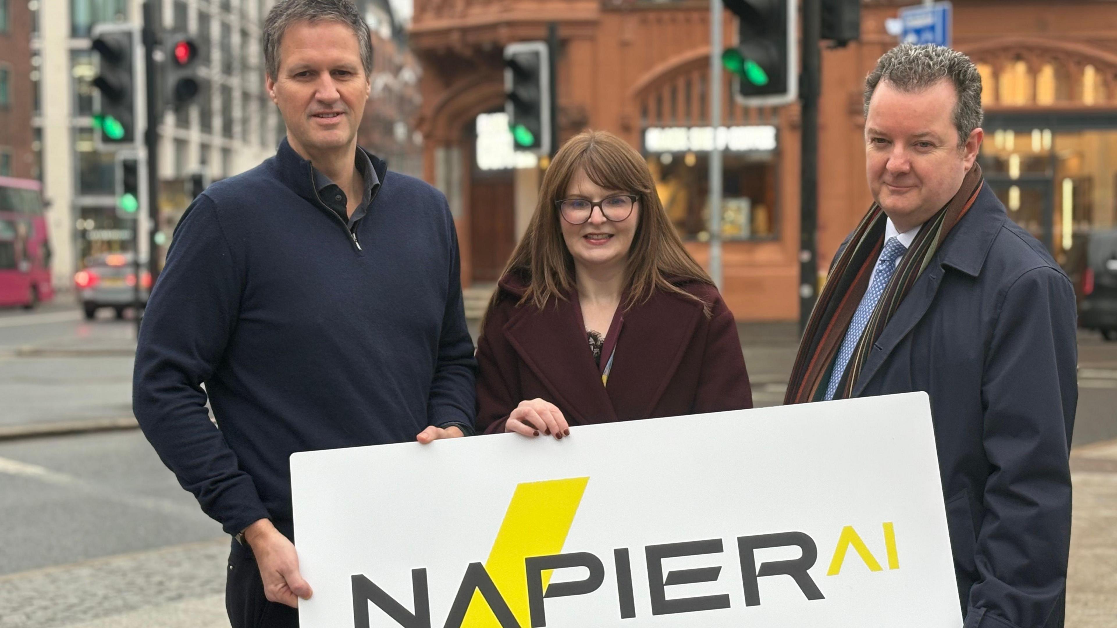 Napier AI's CEO Greg Watson, Economy Minister Caoimhe Archibald and Invest NI CEO Kieran Donoghue stand holding a large white sign with NapierAI written on it. Mr Watson is wearing a navy quarter-zip jumper, Ms Archibald is wearing a burgundy coat and Mr Donoghue is wearing a navy coat and a striped scarf.