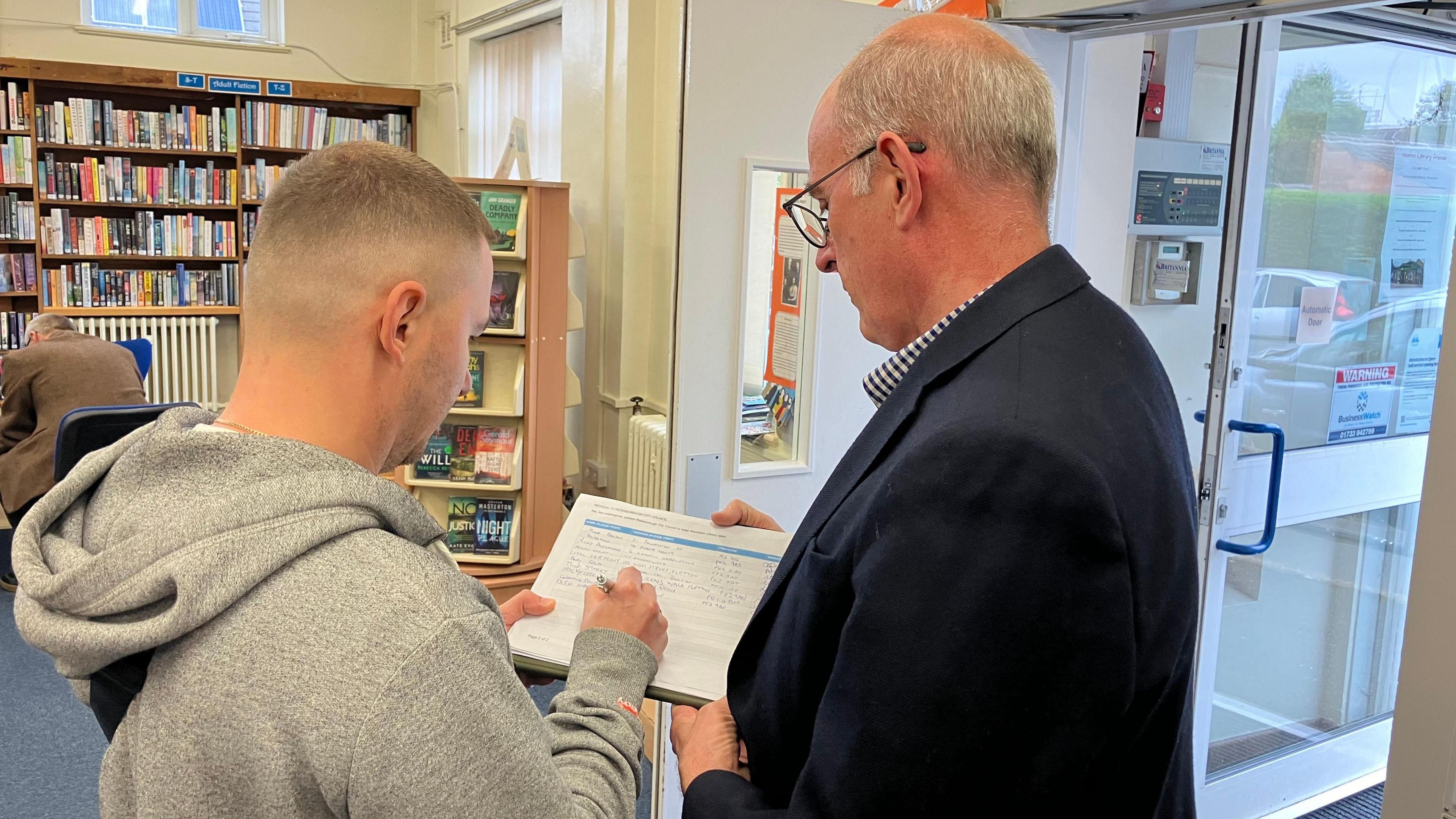 In the library Andy Coles, wearing a black blazer, stands on the right and holds the petition while a man in a grey hooded sweatshirt signs it