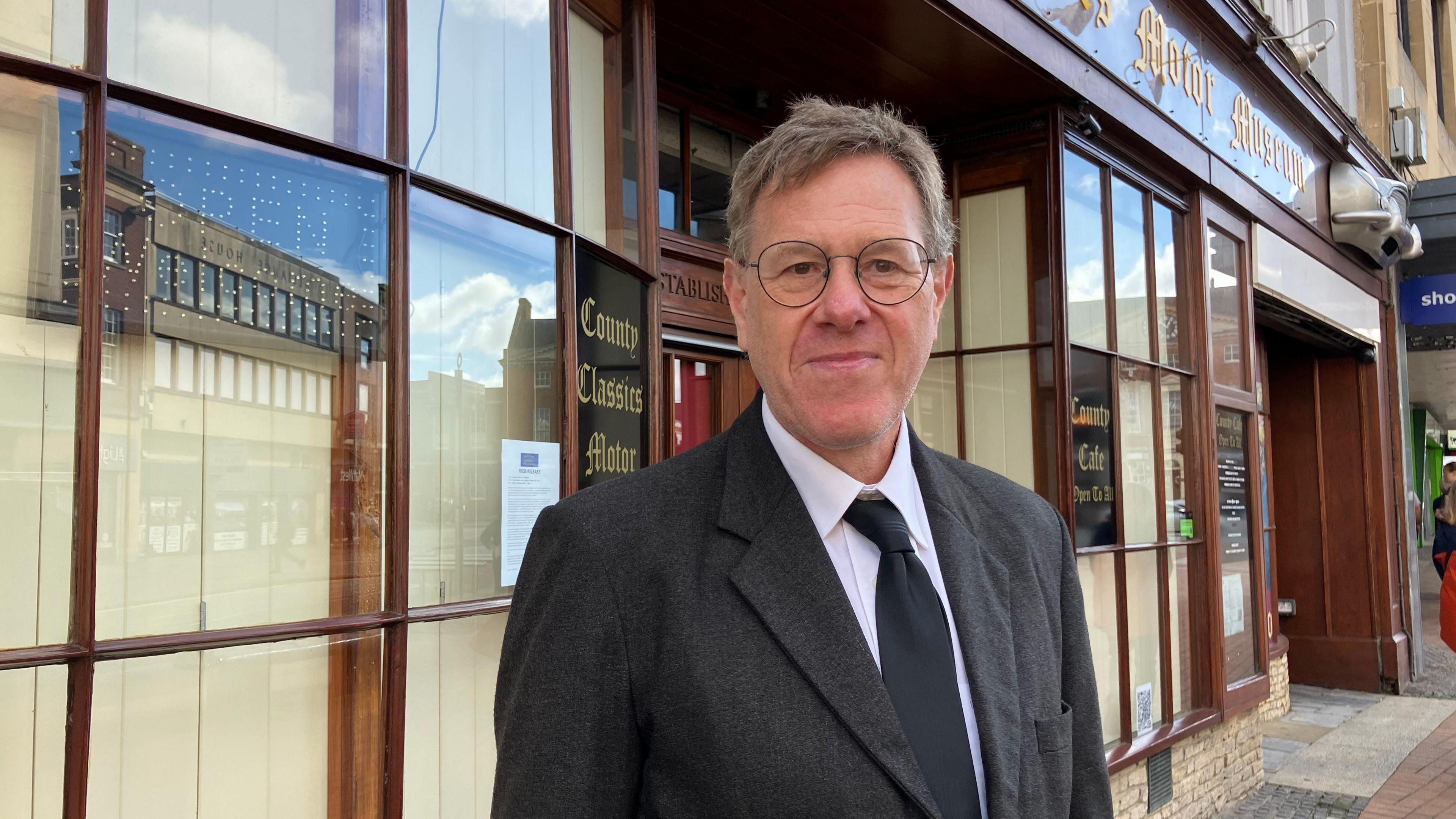 Nick is stood in the centre of the frame, looking at the camera. He is wearing a black suit and tie and has a slight smile. Behind him is the covered windows to the museum.