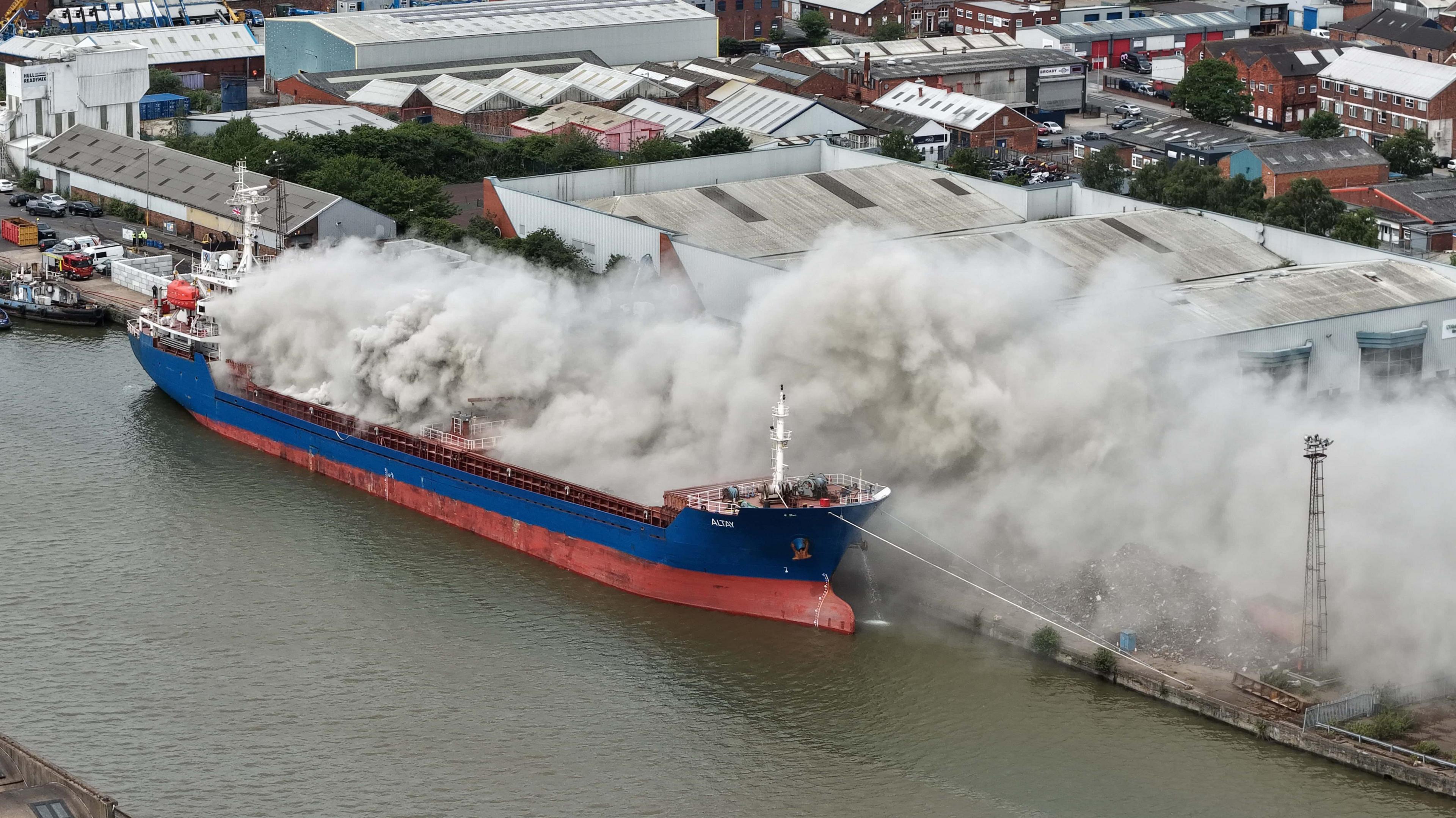 An aerial view of a blue and red vessel with grey smoke billowing out from the deck. A number of industrial buildings are visible in the background.