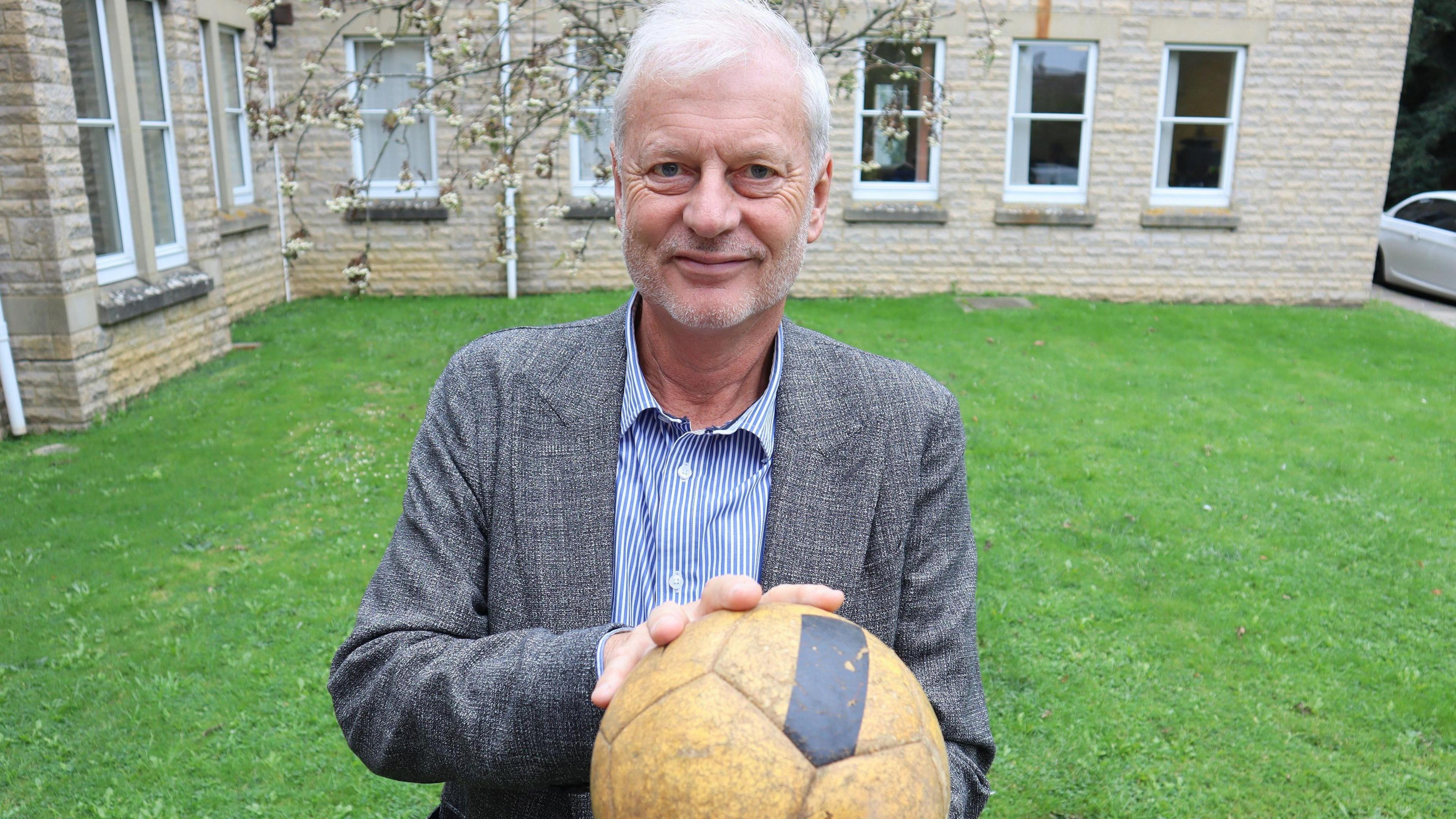 Councillor Tim Sumner stands in a grass area, there is a building in the background with pale beige brickworks and a blossom tree, he stands in the middle of the of the image holding a yellow football with a black stripe. Councillor Sumner has white short hair and is looking directly at the camera, smiling and wearing a blue and white striped shirt with a grey tweed blazer over the top.