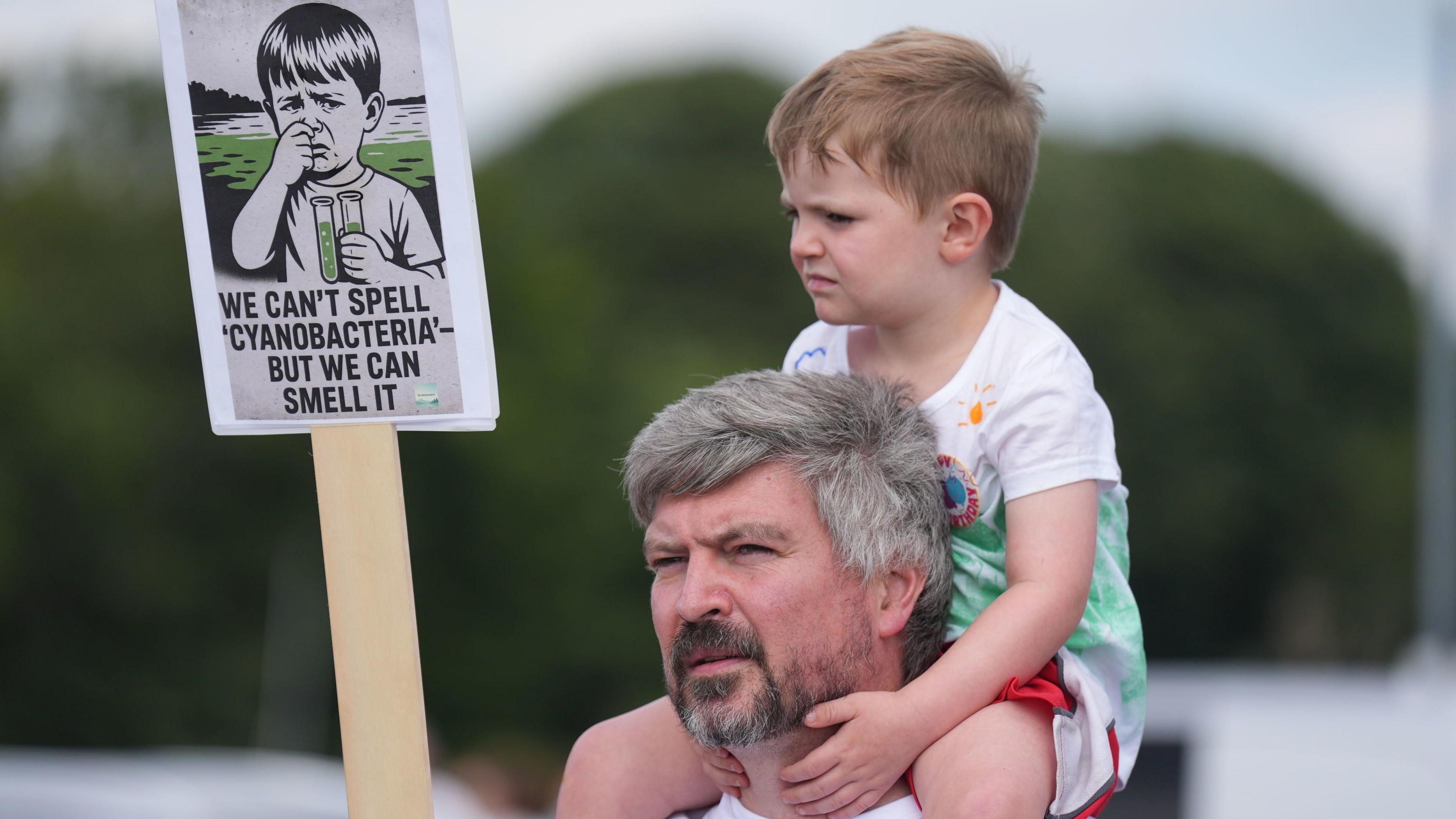A man with grey hair and a grey beard has a young boy perched on his shoulders. The boy is holding a sign, which reads: "We can't spell 'cyanobacteria' - but we can smell it". The sign also depicts a young boy with two test tubes full of green fluid with a sad expression on his face.