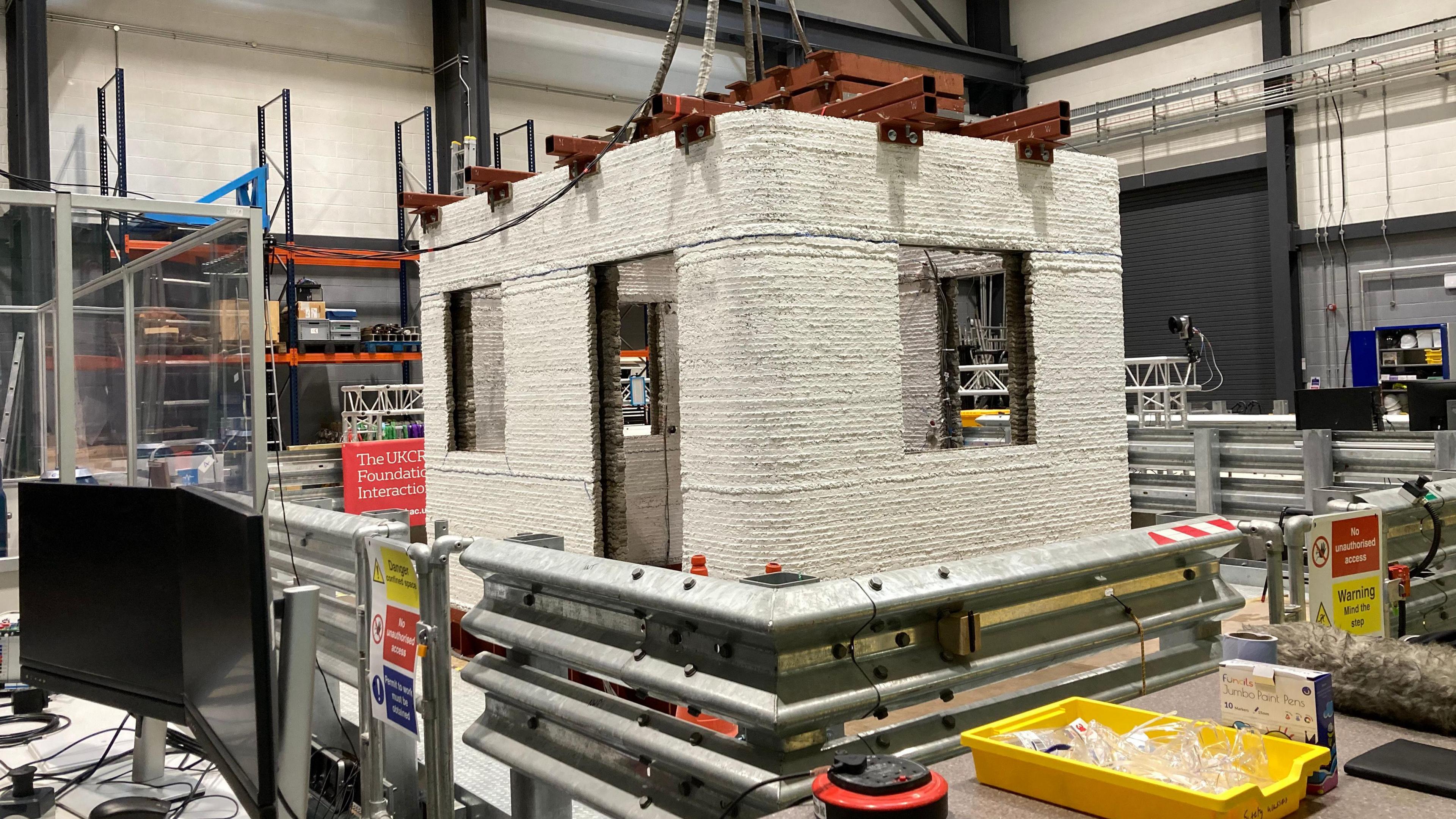 A white 3D printed structure in the shape of a building has been erected in a laboratory. The structure is being held down by steel. There are warning signs and a yellow tray full of protective eyewear.