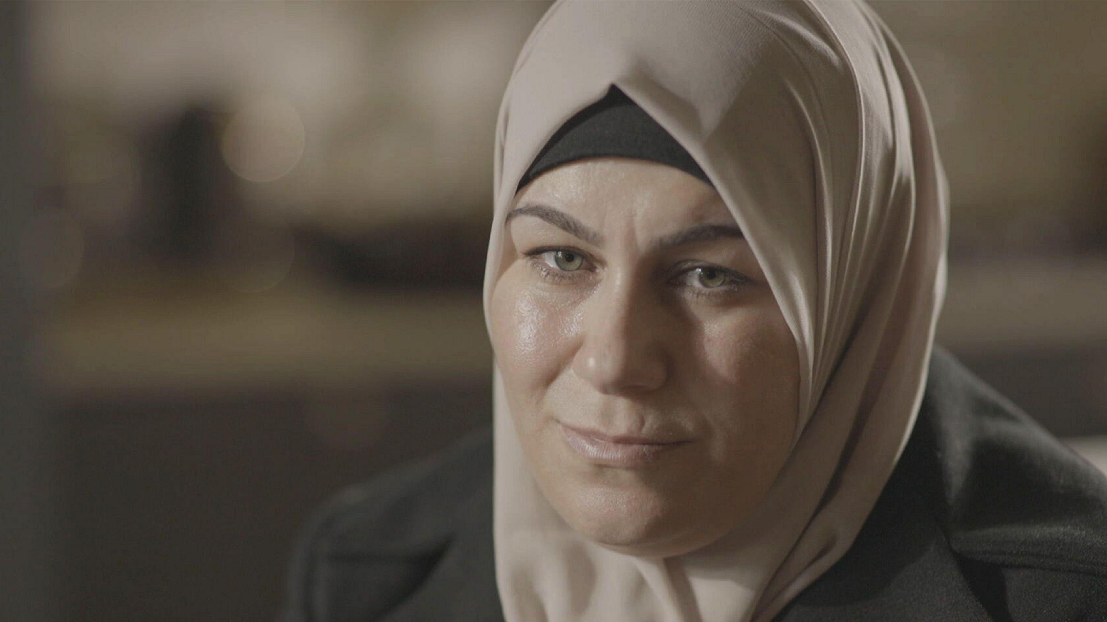 A woman dressed in a headscarf is sitting in a room looking to the side of the camera. The woman has green and hazel coloured eyes. Her headscarf is made of a fine silky material and is beige in colour. She is also wearing a dark coat. Behind her, the room is out of focus with nothing visible.