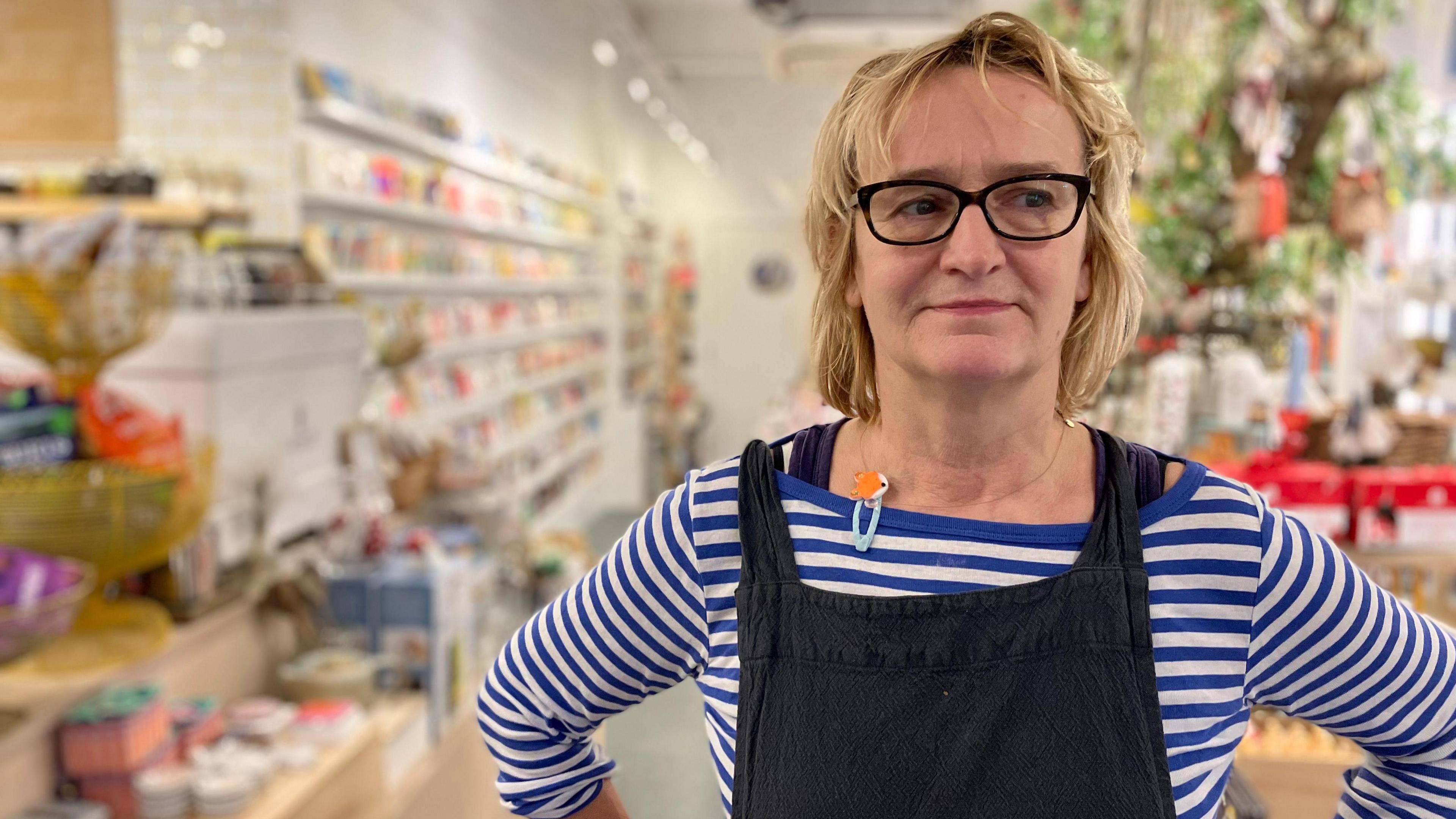 Justine wearing a black apron with a blue and while long sleeve top underneath has blond above shoulder length hair and black rimmed glasses. She is standing with hands on hips in front of the blurred background of her shop.