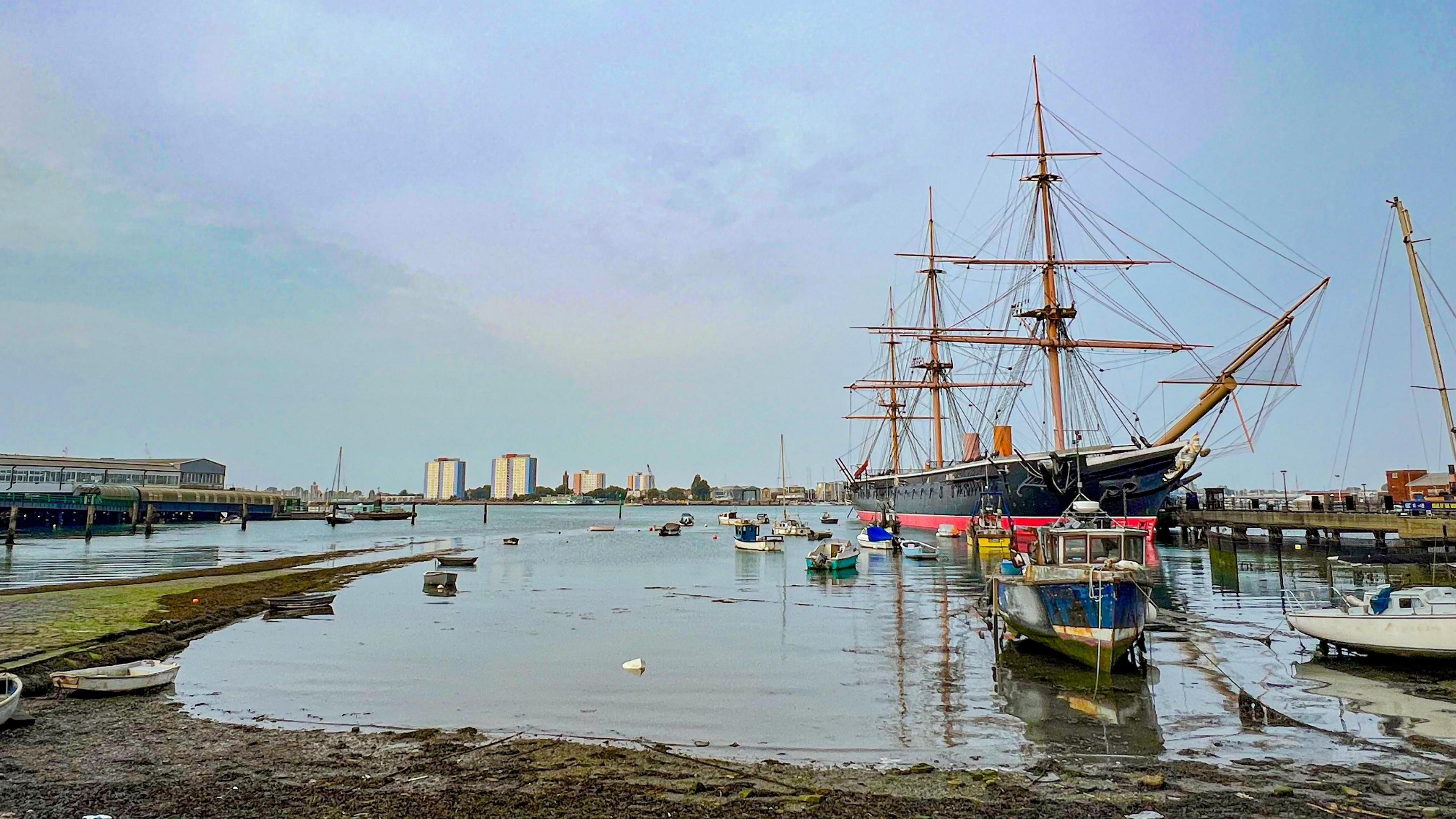 HMS Warrior - a 19th Century battleship - sits in the water at Portsea under blue skies surrounded by several small boats and with buildings seen in the distant background. 