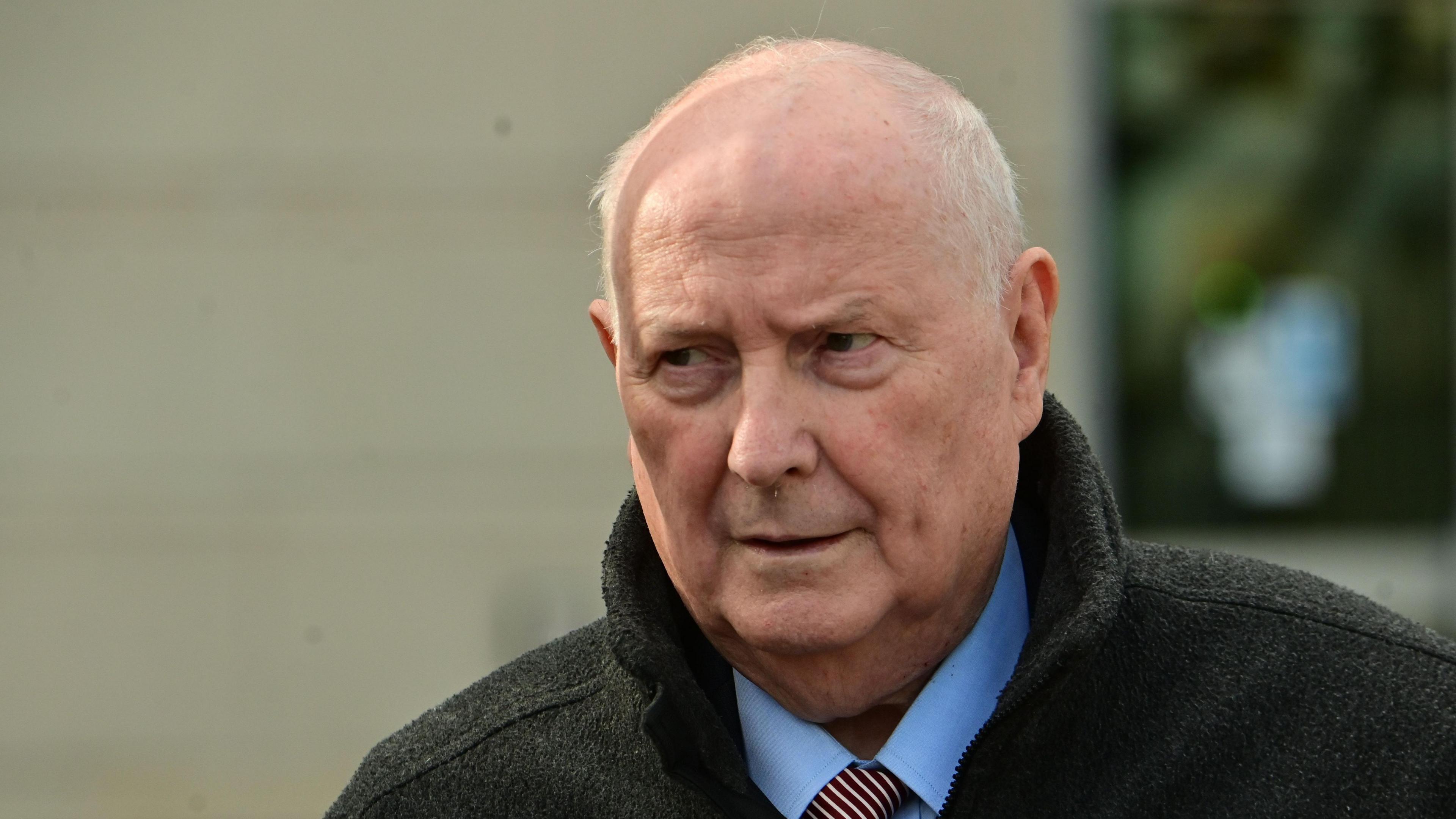 A man with balding white hair is looking off to the side of the camera. He is wearing a dark coloured coat, a blue shirt and a striped tie.
