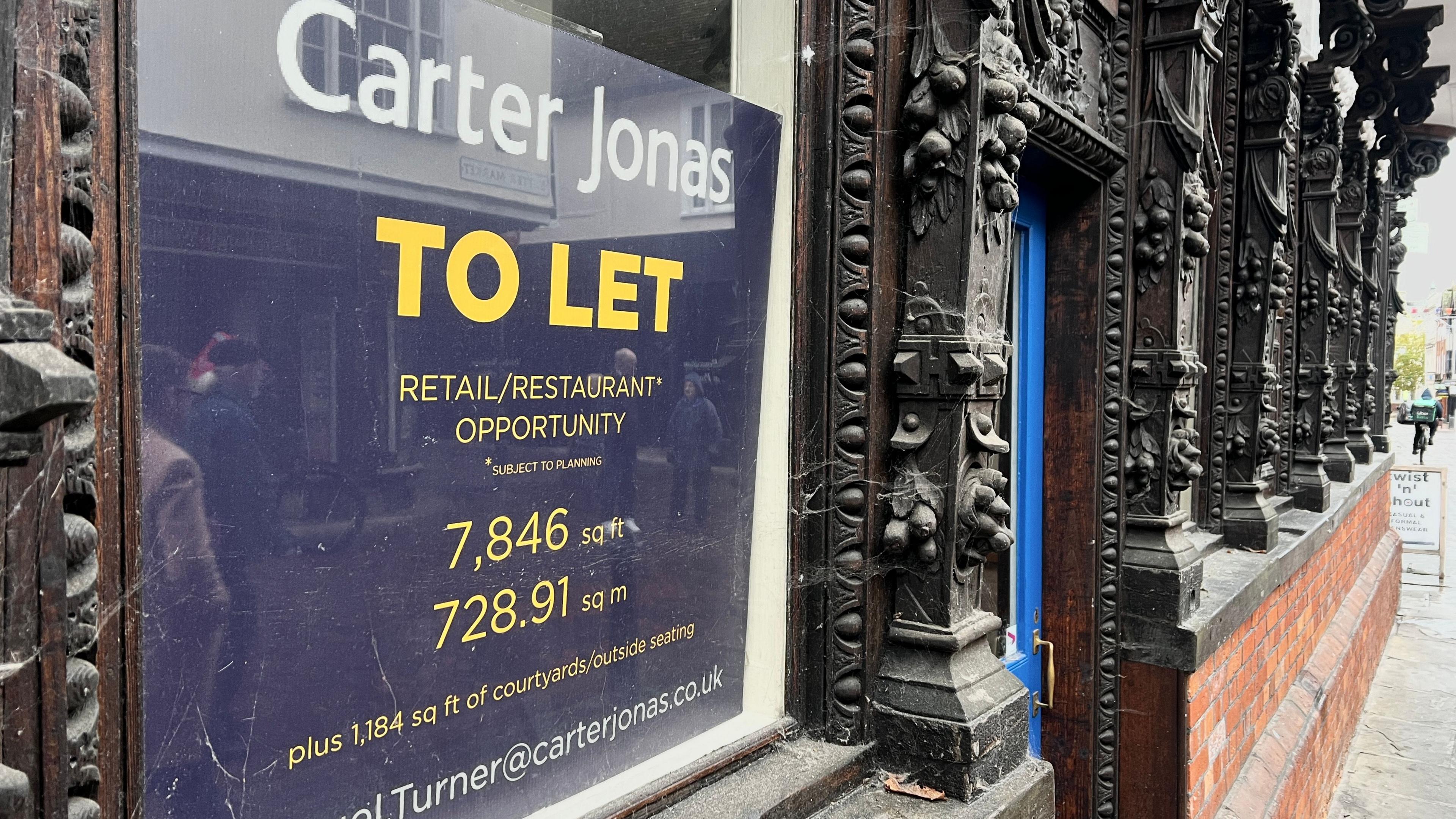 A 'to let' sign sits on a shop front which has been closed. It details the size of the property and how interested parties can get involved. The shop front has black carved details.