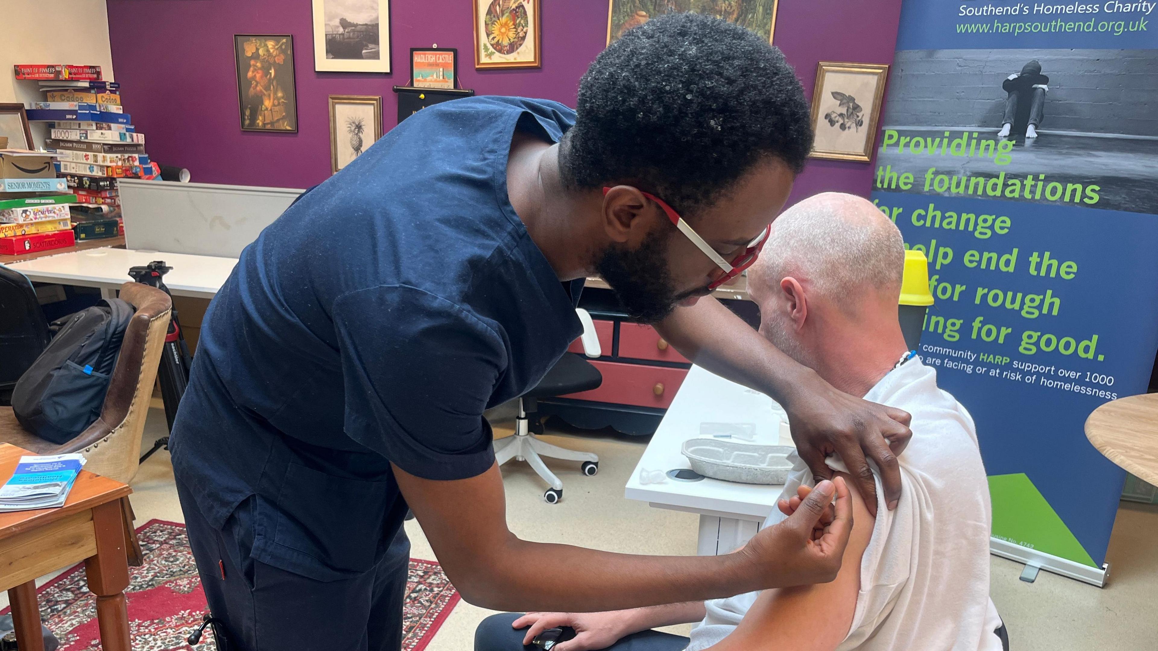 A member of the NHS outreach service gives a vaccination to John, a rough sleeper in Southend. The member of staff is in the main games room in the HARP hostel. He has a table beside him and is wearing dark blue scrubs. John is wearing a while T-shirt with the sleeves pulled up so he can be vaccinated.