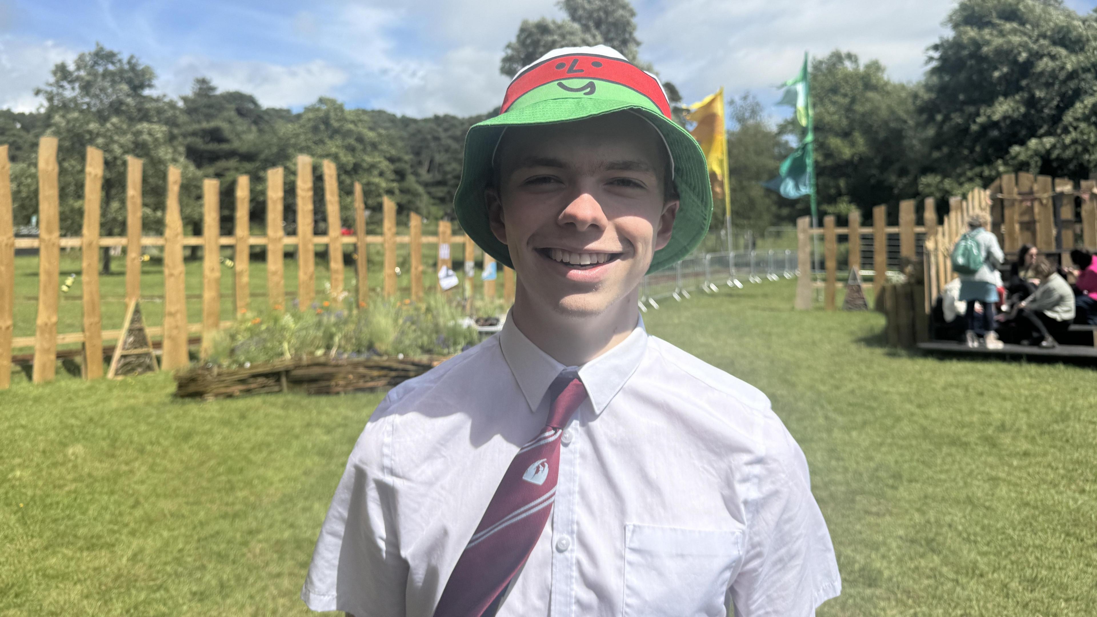Cai, wearing a white shirt, burgundy tie with red stripes, and a white, red, and green striped bucket hat with Mistar Urdd's face on it. He is standing on the Maes, with grass and wooden fencing behind him.
