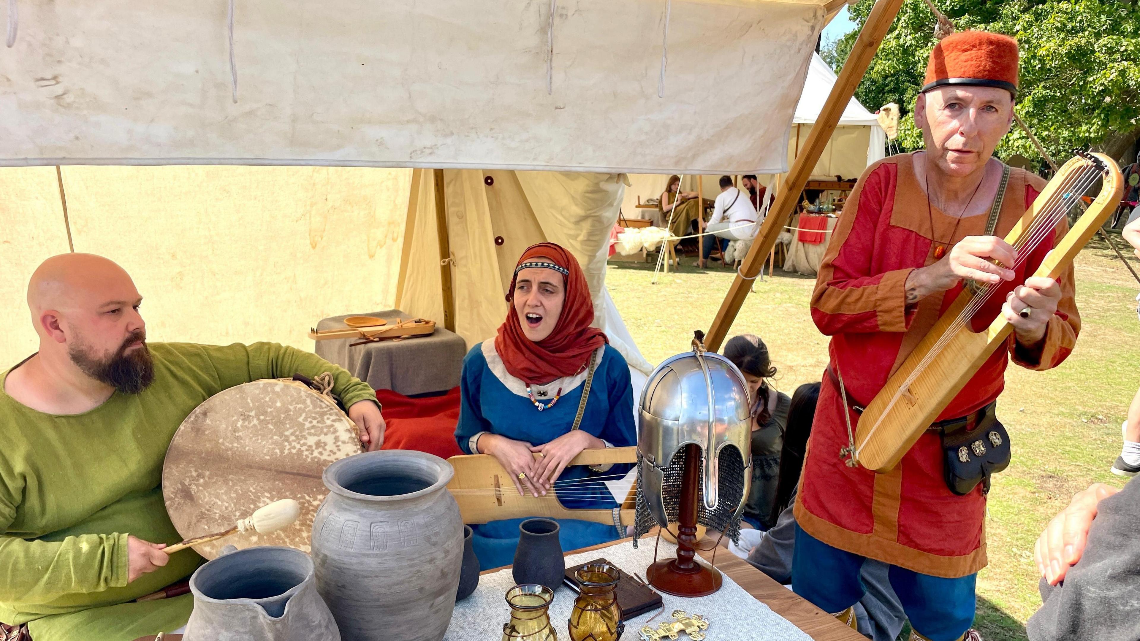 Three reenactors including two men and a woman are dressed like they are from Anglo-Saxon times and they play instruments and sing.