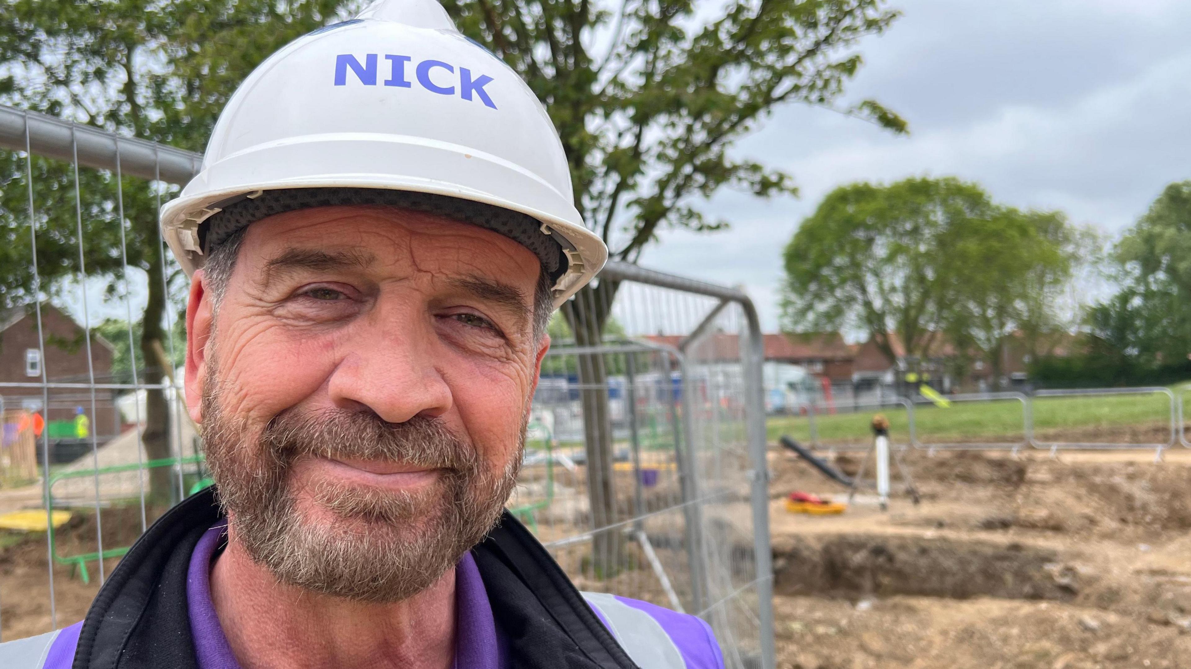 TV presenter Nick Knowles smiles into the camera. He has a beard and is wearing a white hard hat, purple t-shirt, black top and a purple high-vis vest. He is standing in front of a building site and a metal fence. Trees and houses are visible in the distance.