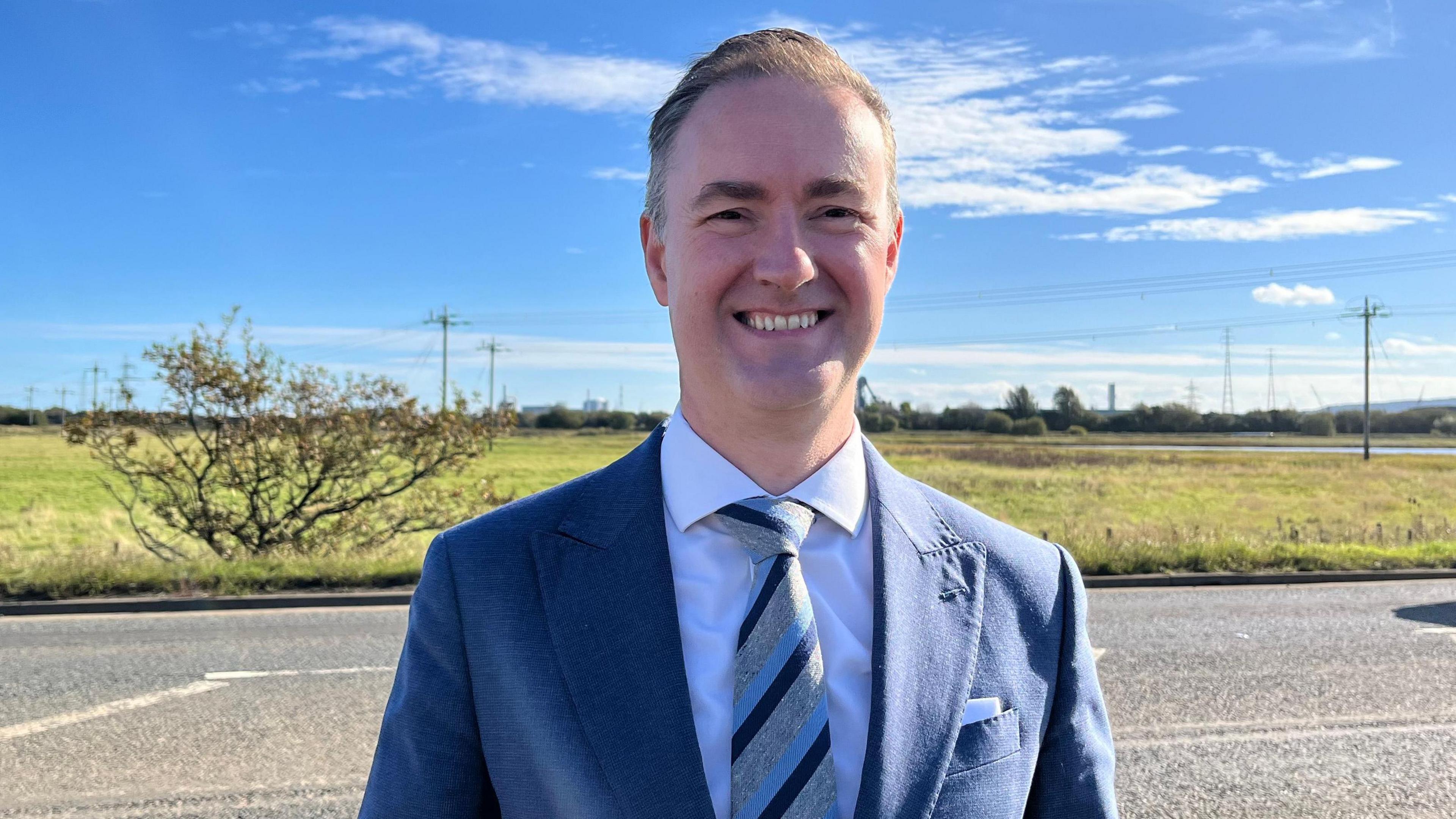 A man with grey swept over hair, a white shirt and blue and grey tie and navy blue suit on, standing in front of a road on a sunny day with green space behind the road. He is smiling.