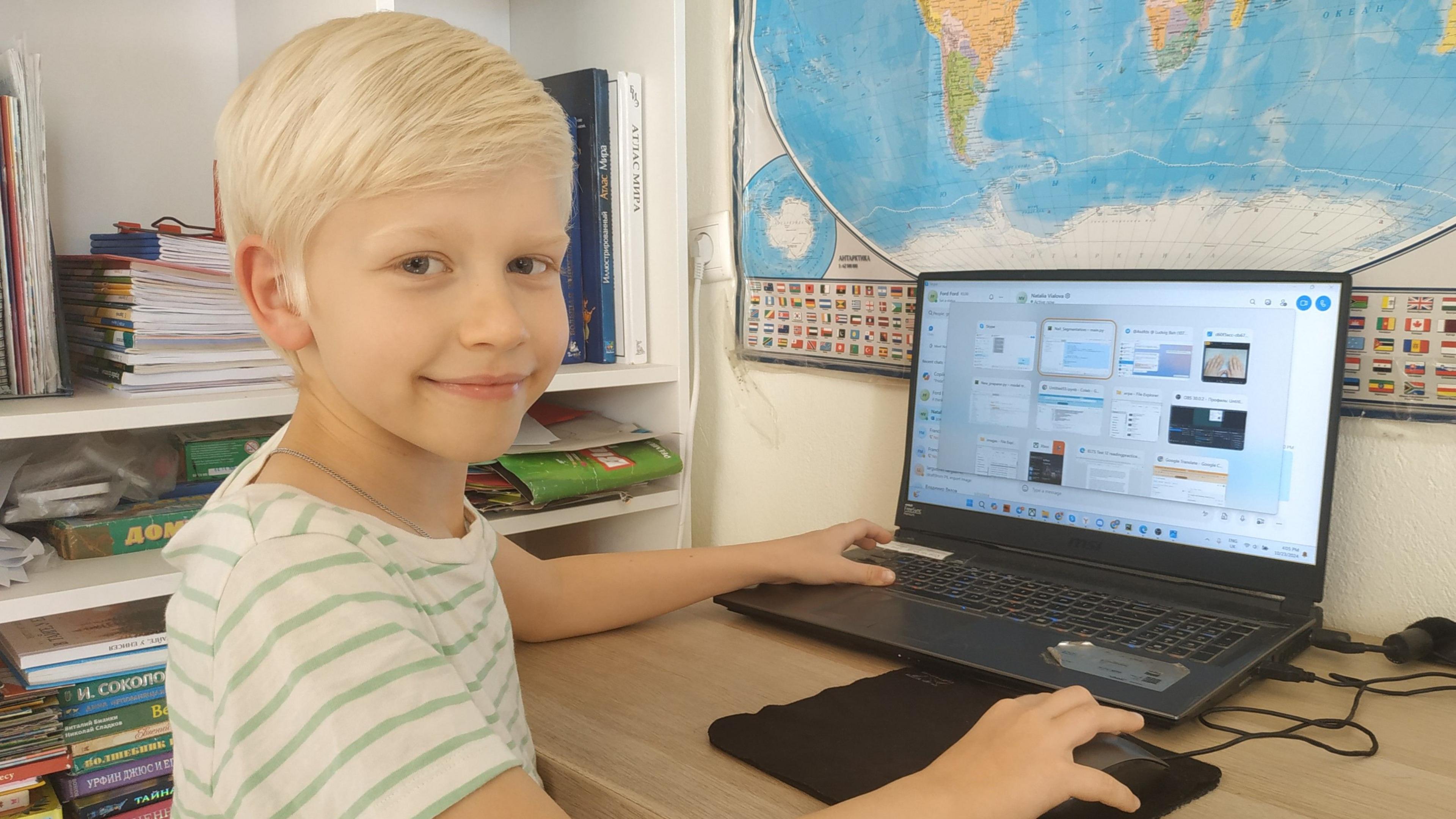 Blonde-haired seven-year-old Sergey sits with his laptop at his desk, smiling at the camera with one hand on his mouse. Bookshelves are seen behind him and world map is on the wall