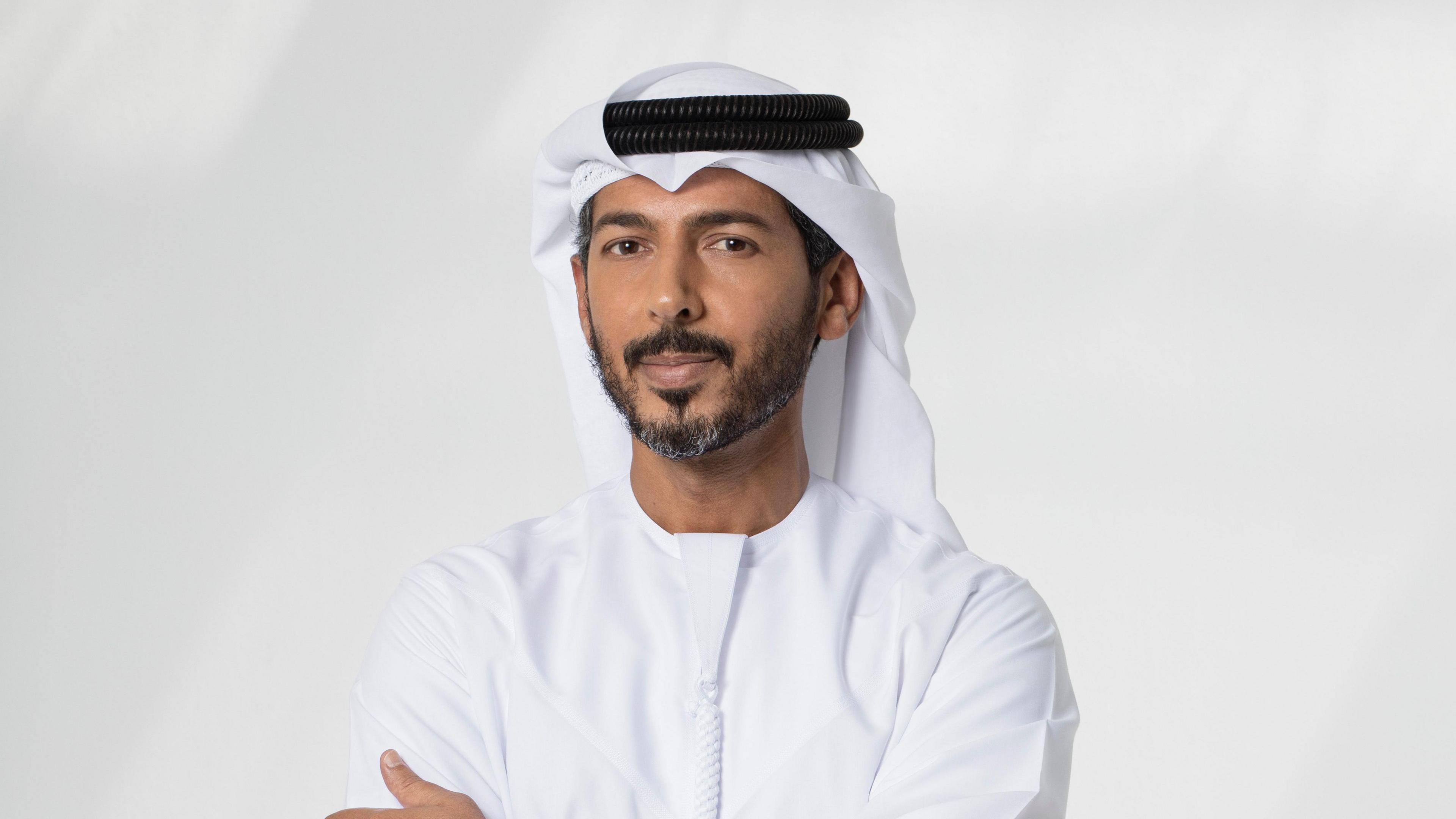 Hassan Alnaqbi Hassan looks at the camera with his arms crossed. He is wearing the traditional emirati dress - a Kandura - a long white robe.
On his head he wears a white headdress called ghutra and secured in place by a black cord called the agal.

