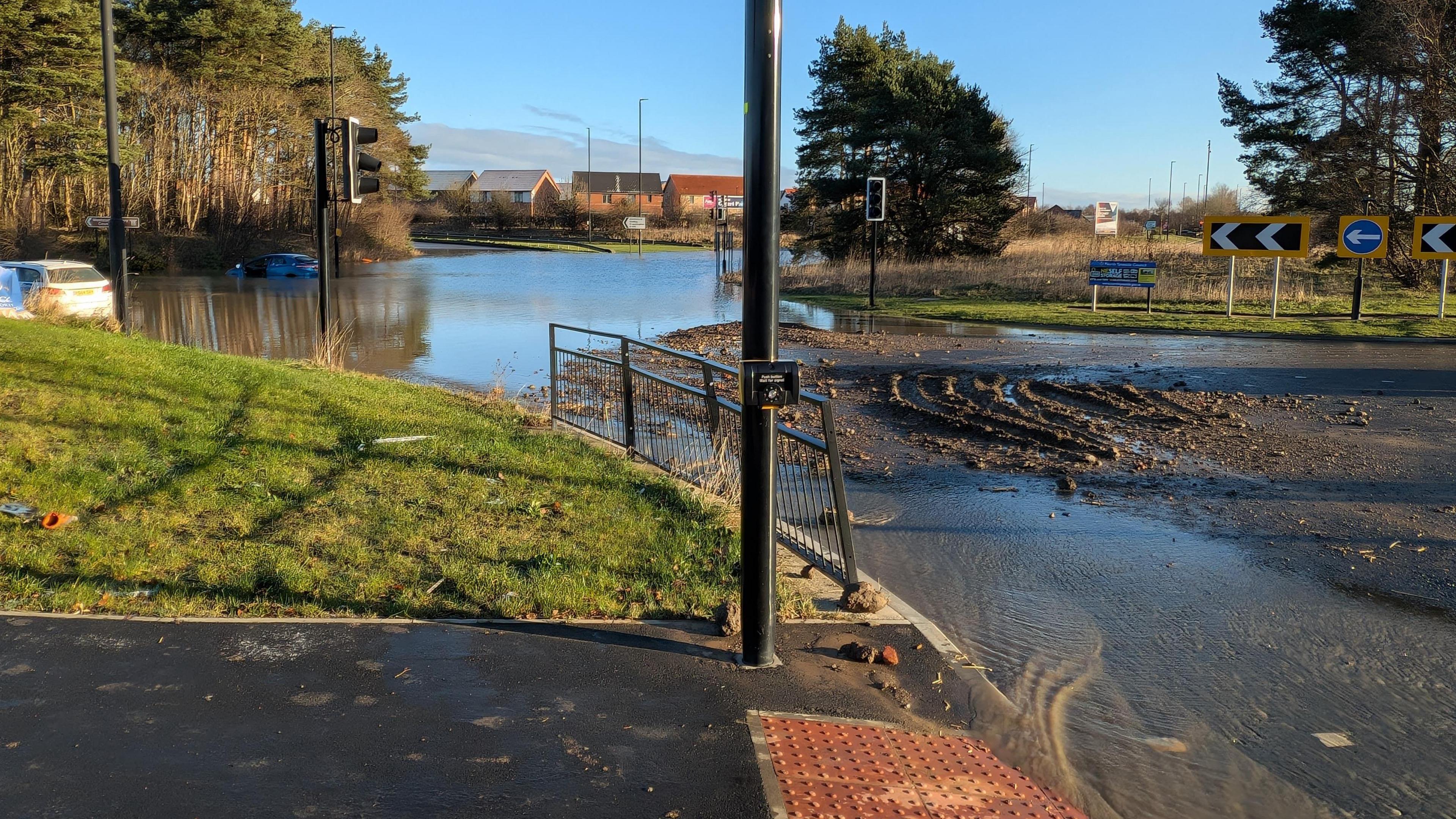 Flooding on the roundabout. Two cars are stuck in the water to the left. To the right, mud and silt has covered much of the road. Tire tracks have been left behind in the mud.