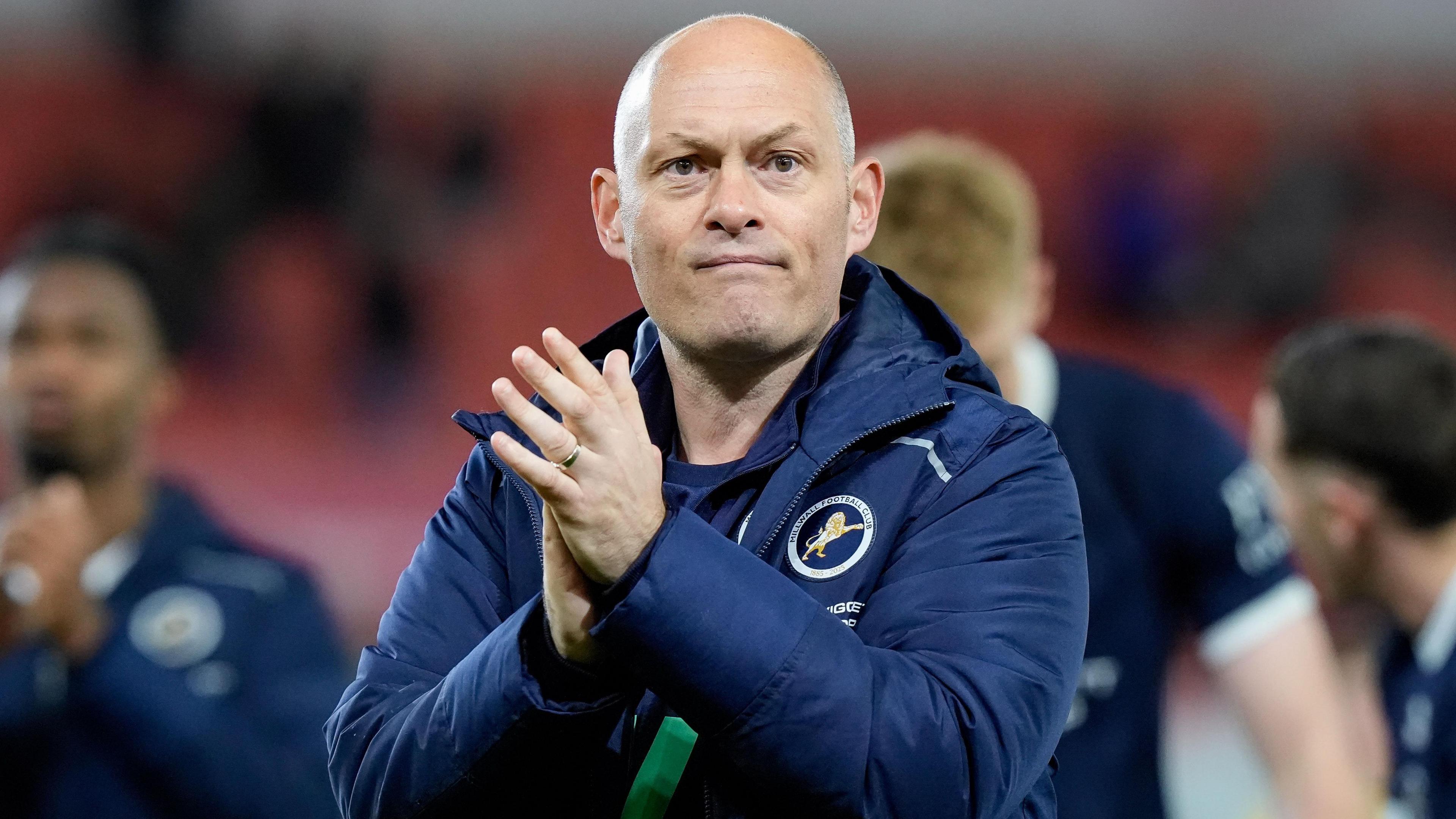 Alex Neil, wearing a navy blue Millwall jacket, claps his hands to applaud the fans after a win against Stoke