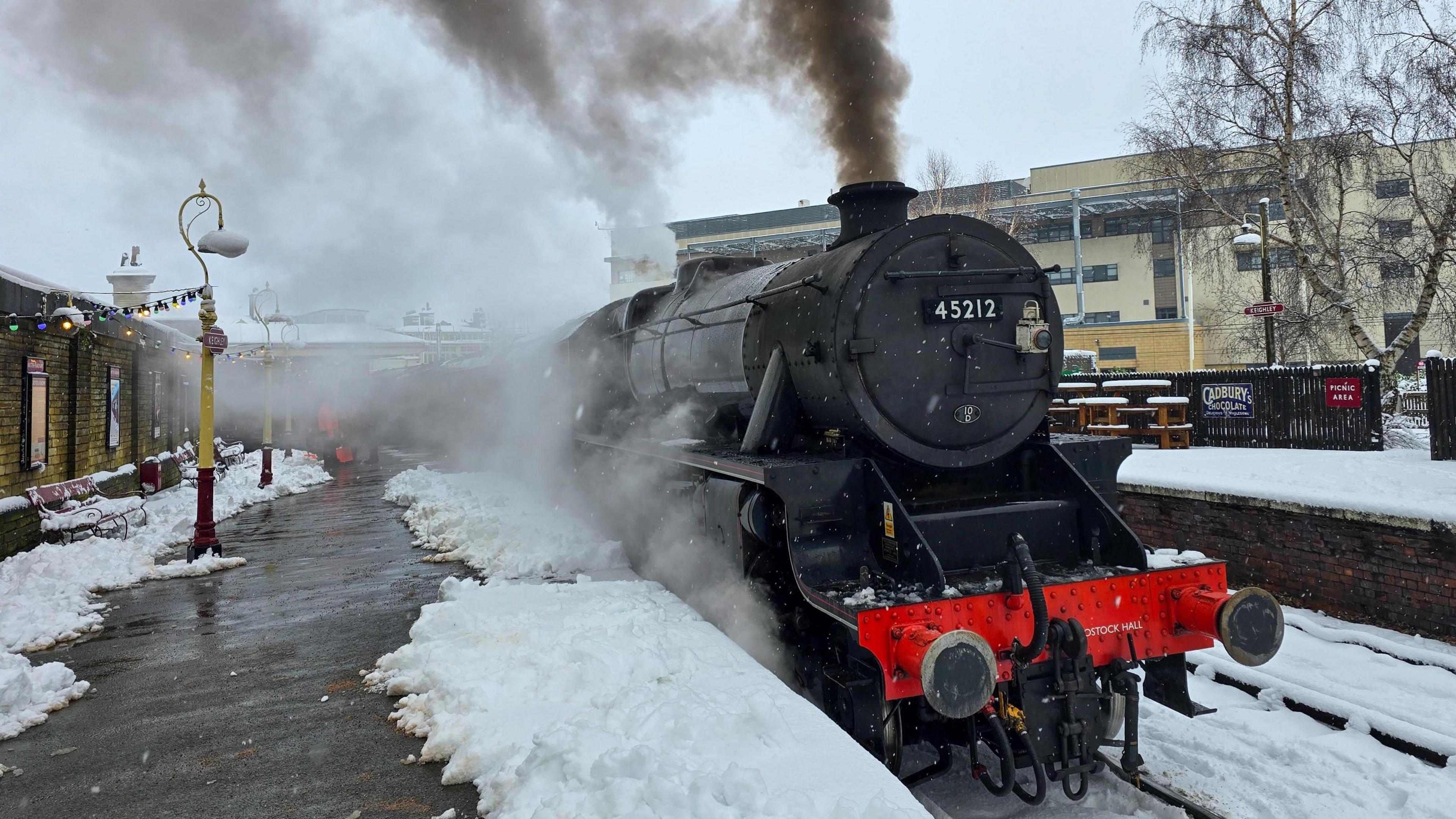 A steam train sits in a station with snow on the platforms. Black smoke comes out of its chimney and steam from underneath it.