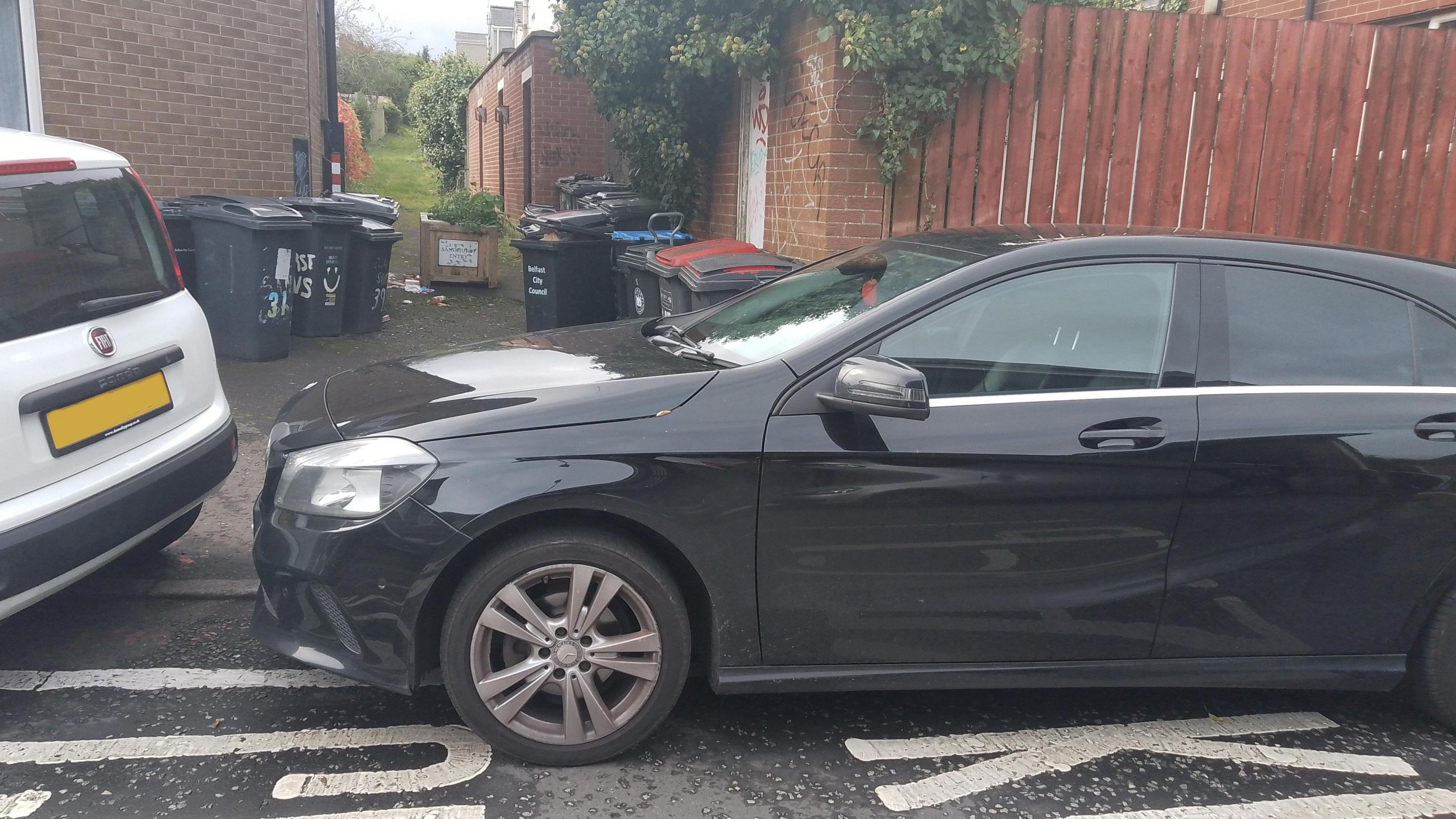 A black car parked on a keep clear zone with an alleyway of bins in the background 