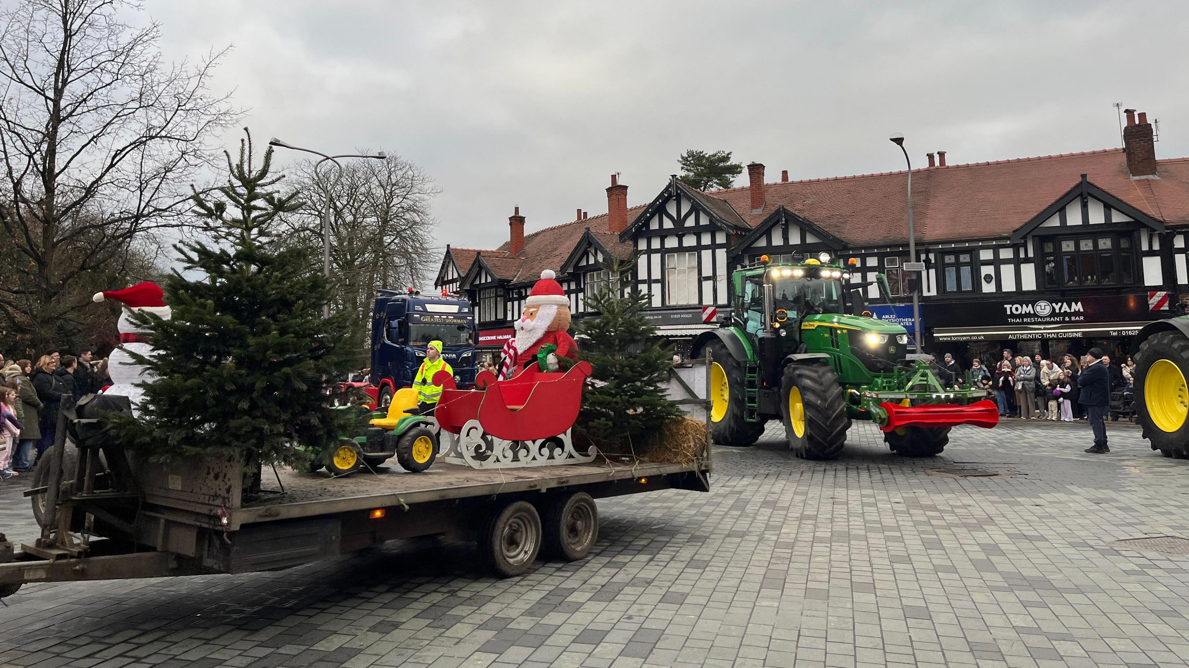 A Santa Christmas display on a cart pulled by a tractor in a town square, with other tractors following behind 