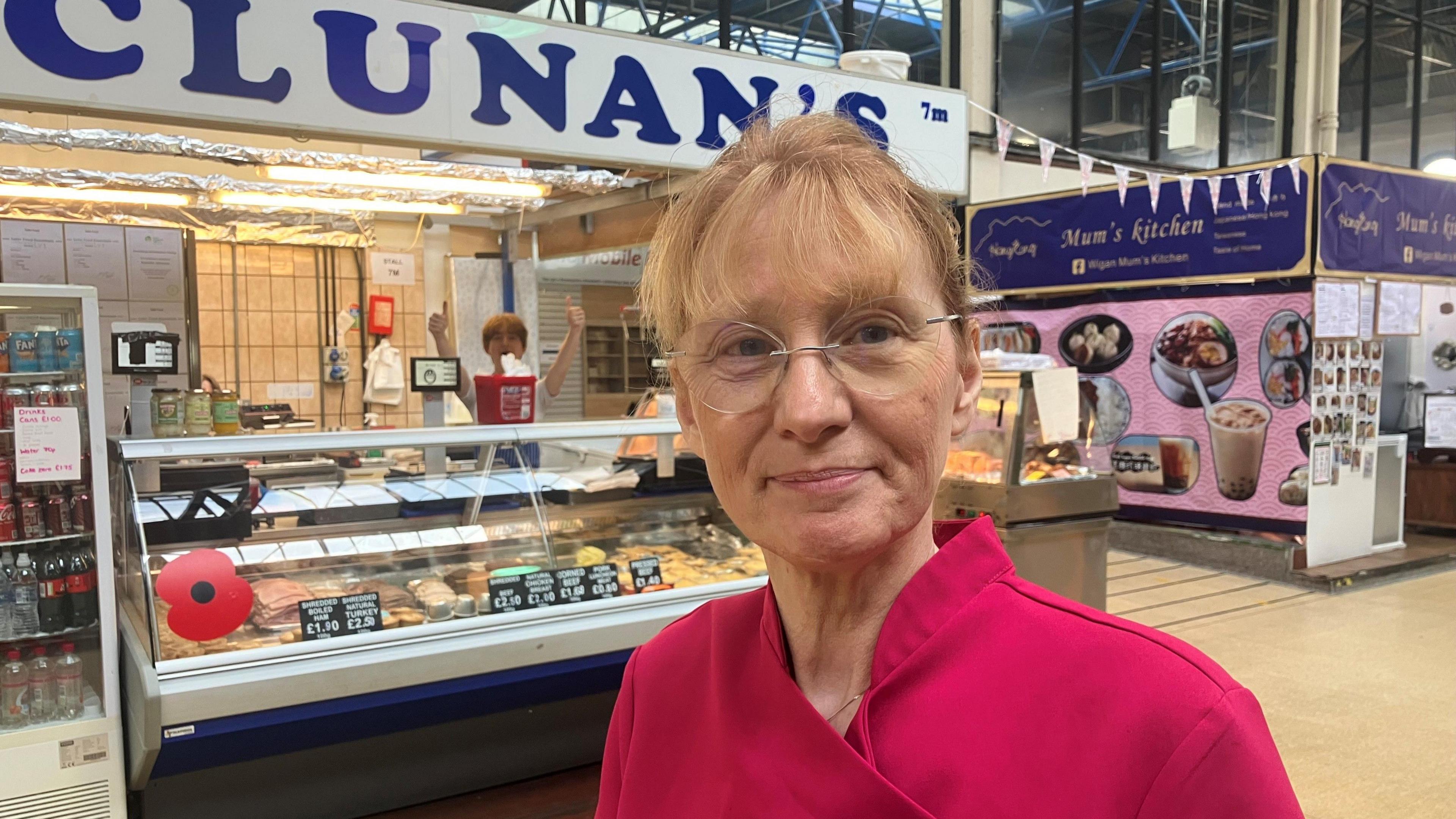 Photograph of Kathleen Clunan from Wigan indoor market. With medium-length, straight blonde hair and glasses, she is pictured wearing a pink top in front of her delicatessen stall.