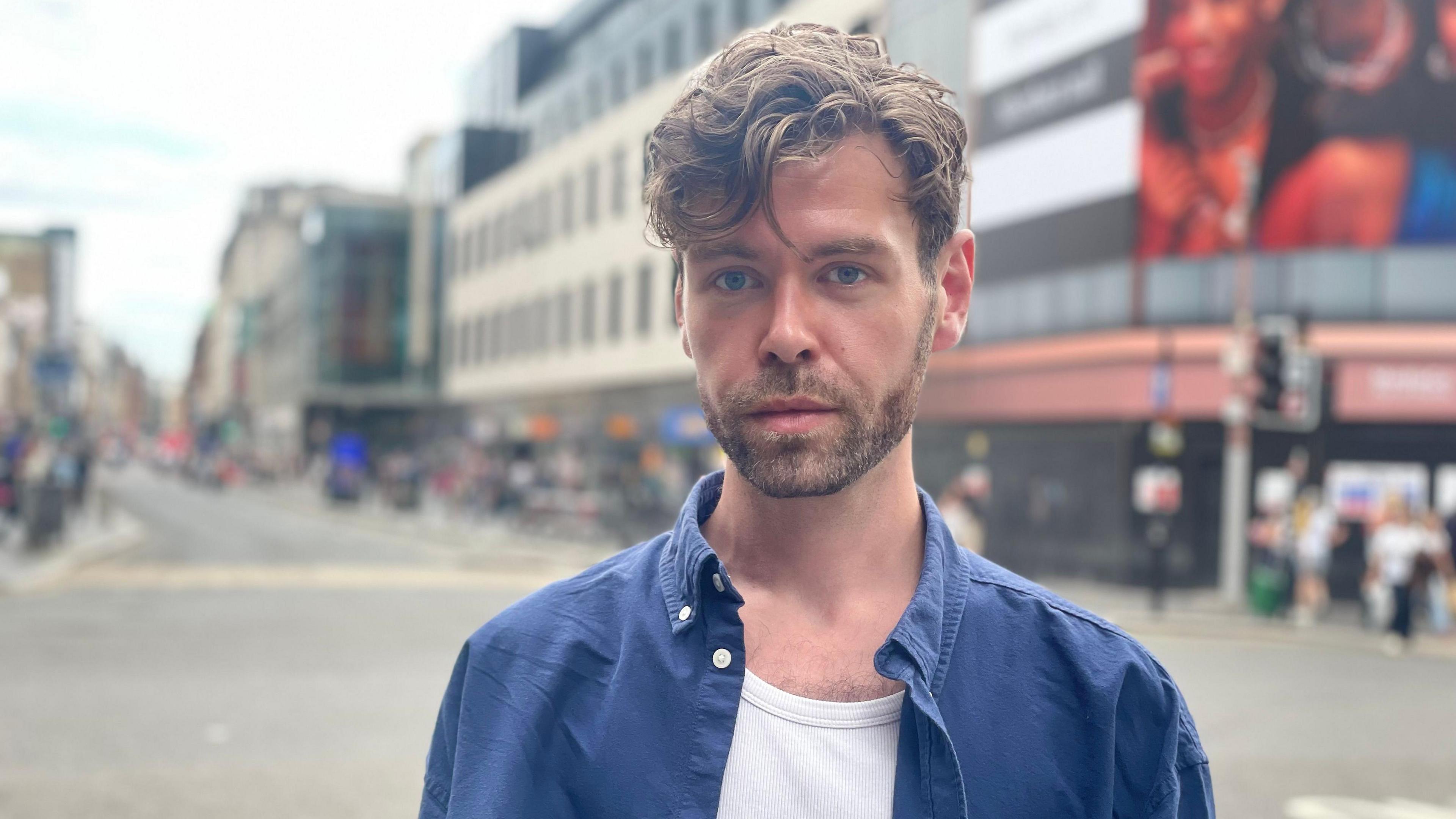 Portrait of Kip Lawson standing on a city street, wearing a blue button-up shirt over a white vest. The background is softly blurred, showing buildings, shopfronts, pedestrians, and a road intersection.
