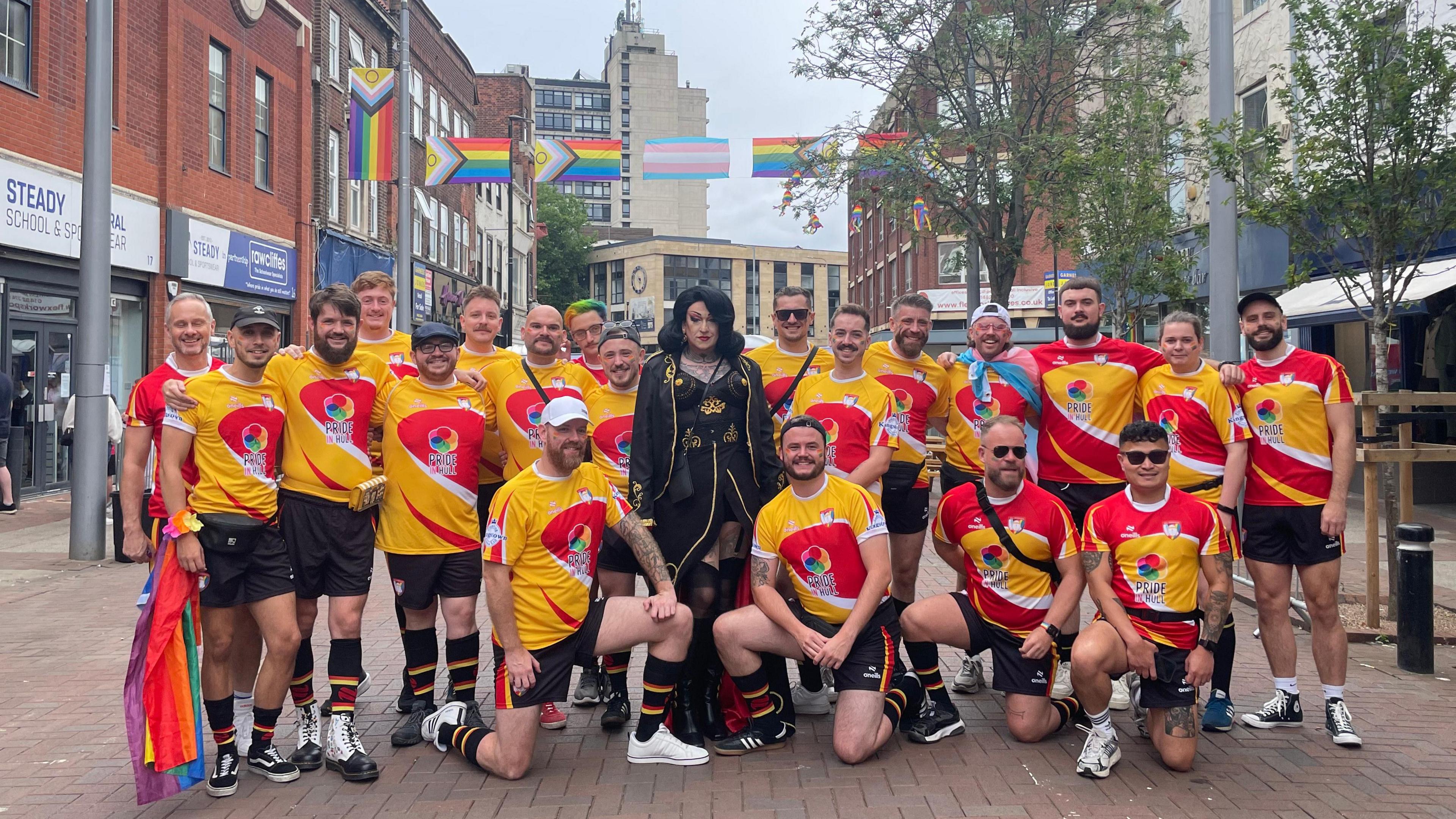 Rugby players pose for a team photo. They are wear red and yellow rugby shirts with a motif on them. A drag queen, all in black with a black 60s styled wig, stands like Cruella de Vil in the middle of them.