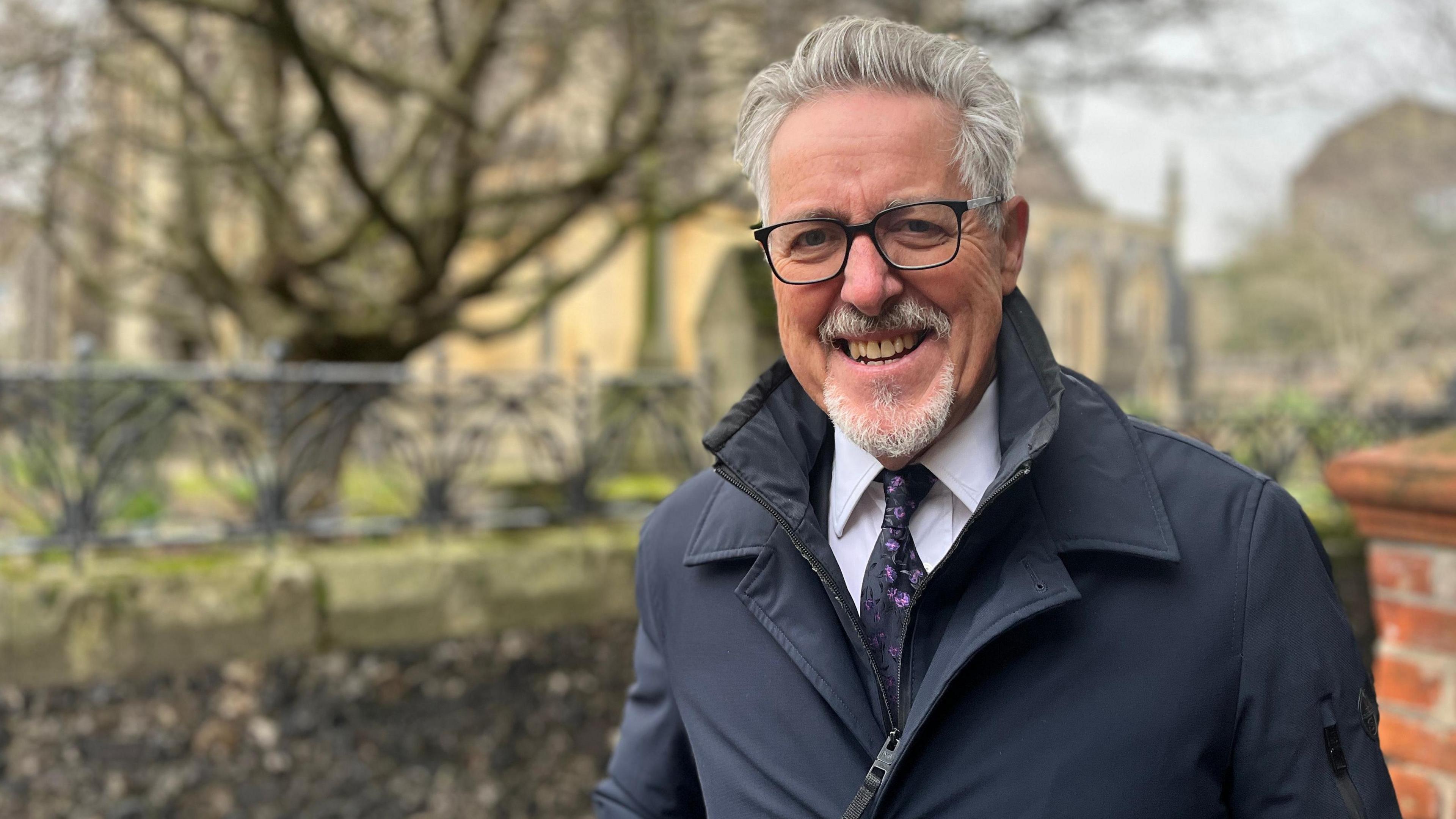 Griff Rhys Jones stands outside Ipswich Minster church yard. He is wearing a navy coat over the top of his suit and tie. He has grey slicked back hair and black framed glasses.