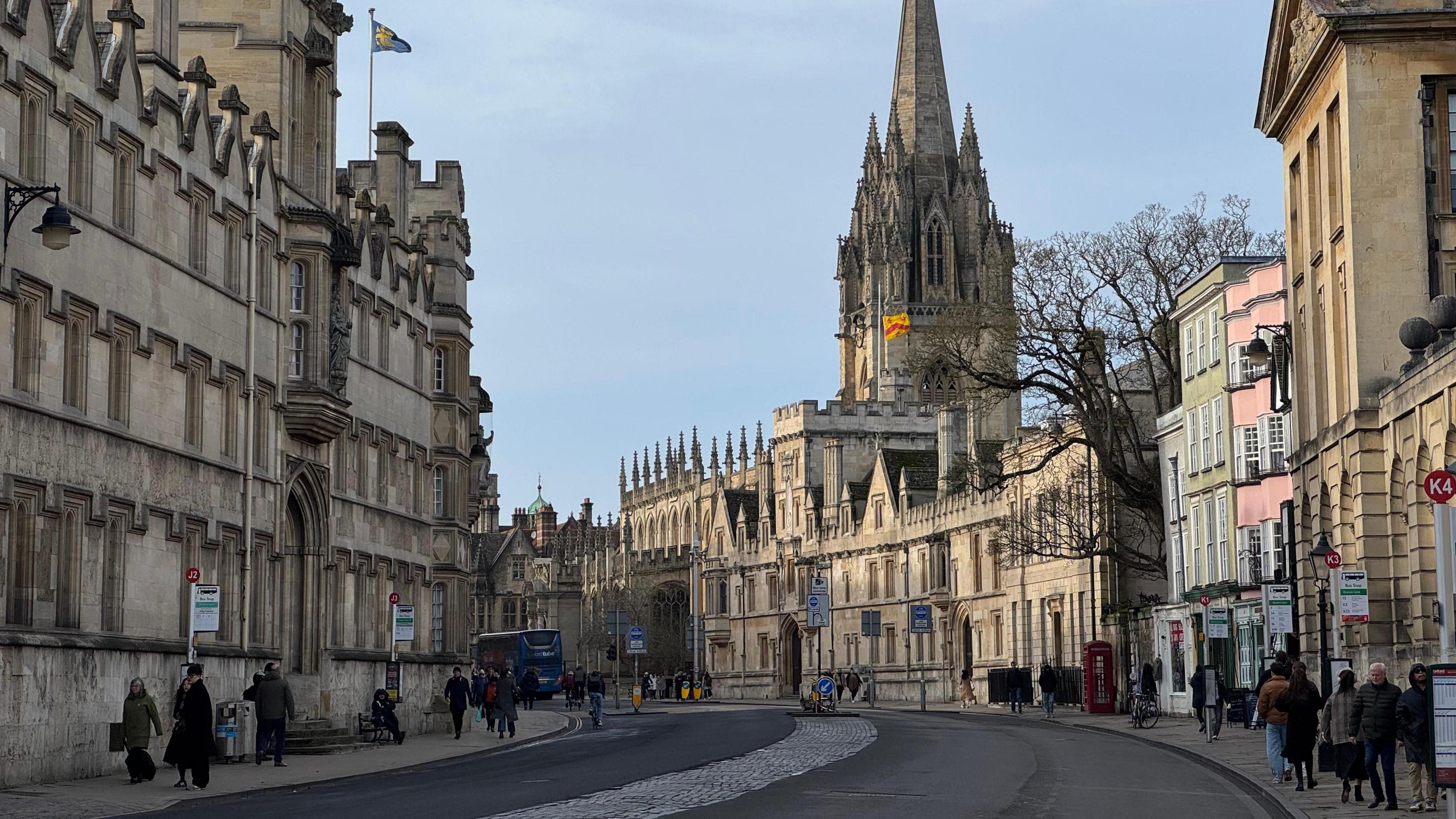 An Oxford street is busy with shoppers. Stone buildings line each side of the road and there is a blue bus. It is a sunny day. In the centre of the picture is a building with a tall stone spire.