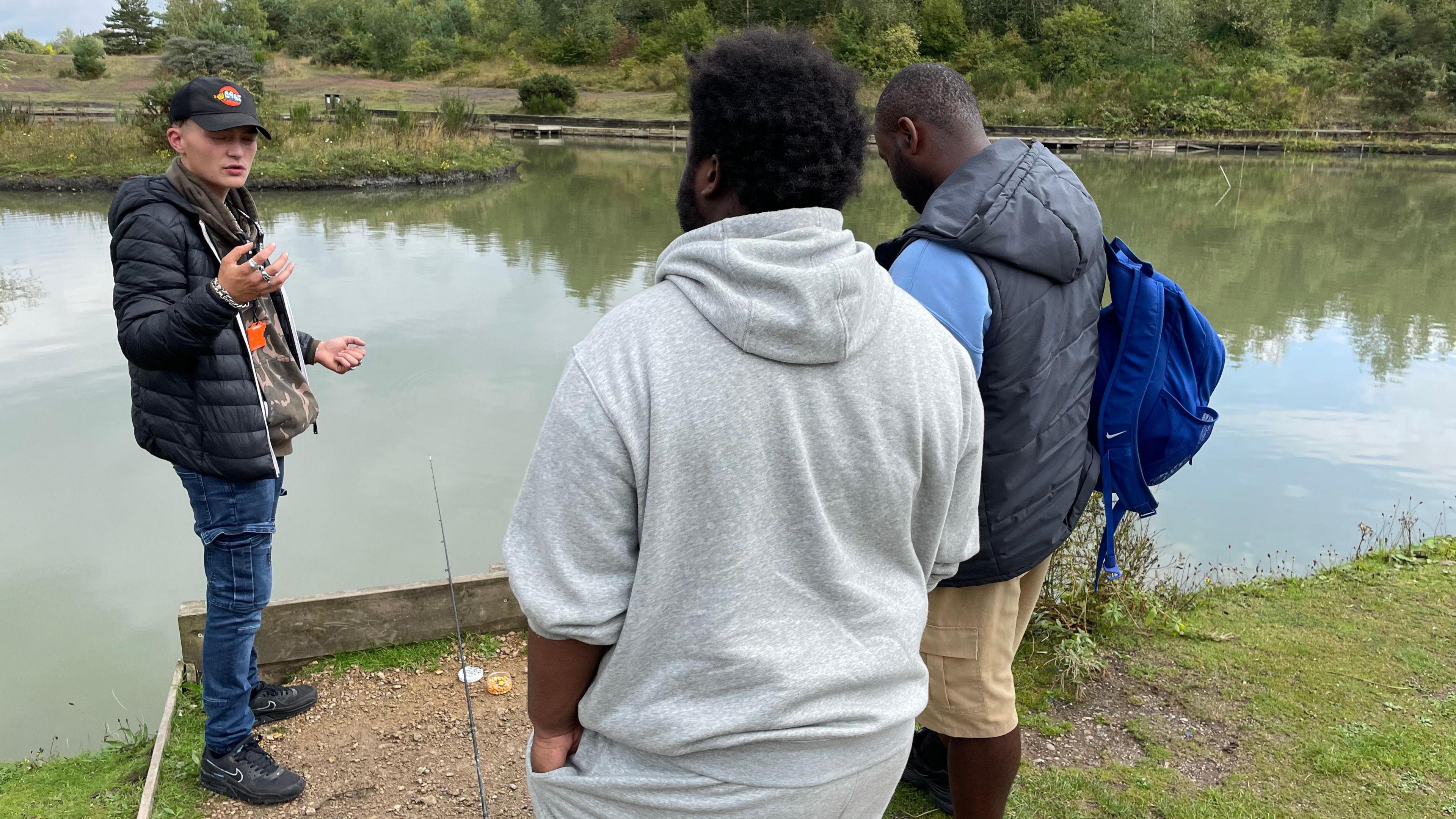 Nico Blade-Smith, wearing blue jeans, a black coat and a black baseball cap featuring the Cast logo, stands at the edge of a fishing lake talking to two men with their backs to the camera. One is wearing a grey hoody, the other is wearing a black gilet  over a light blue top and is carrying a darker blue bag