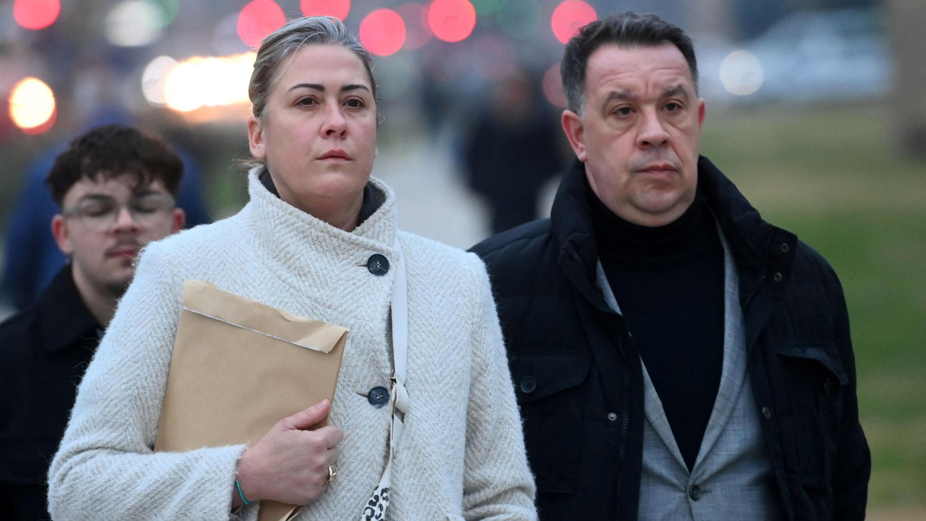 Caroline Darian, a woman with her blonde hair pulled back and a white herringbone wool coat, clutching a manila envelope, and David Pelicot, a man with dark hair and a dark wool coat over a mid-grey suit, walk outside the courthouse in Avignon in December last year, with a determined expression on their faces